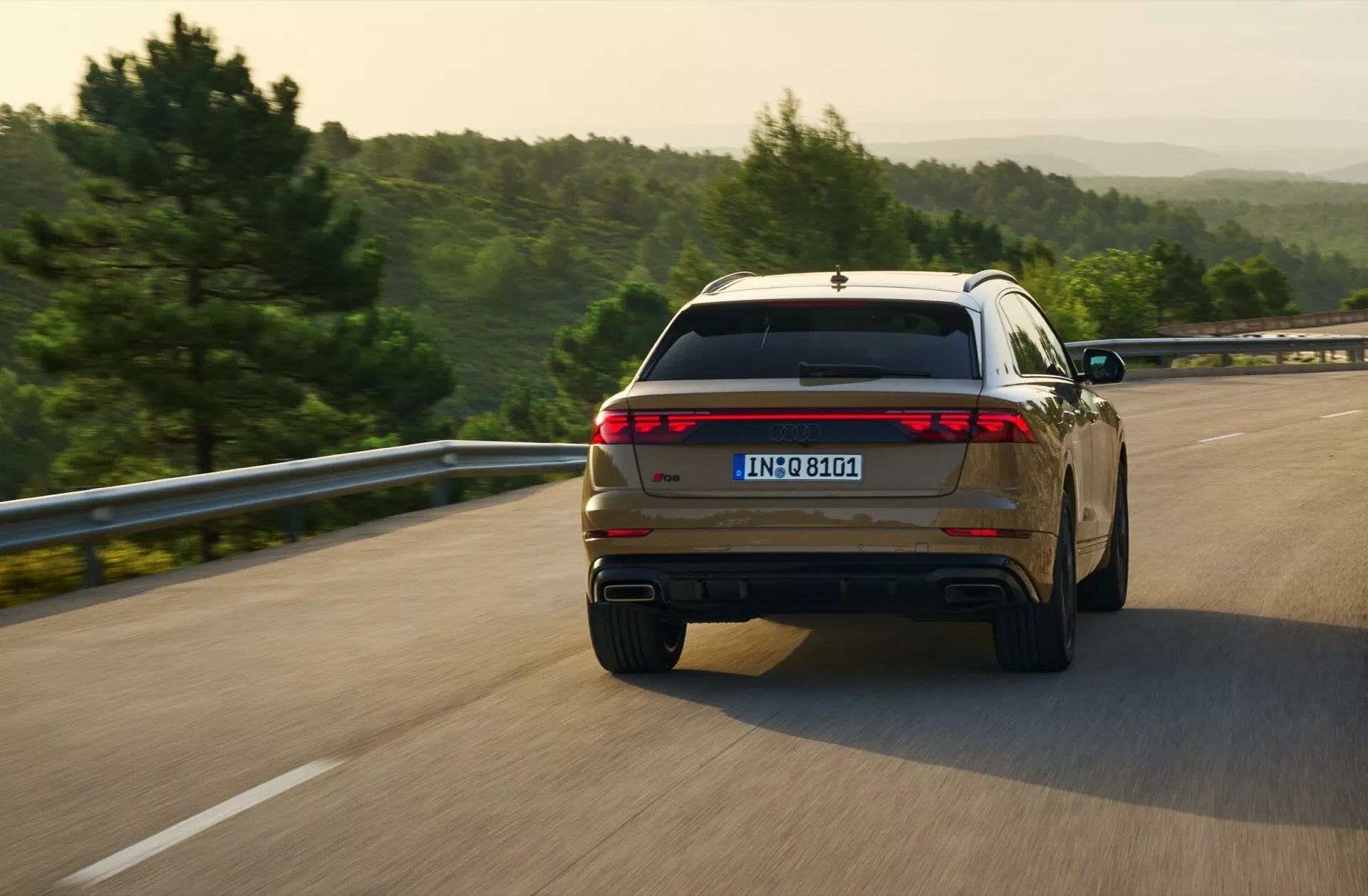 Audi Q8 2023 (4M-Facelift) zachytené v dynamickom zadnom trojštvrťovom pohľade počas jazdy po otvorenej ceste, predvádza jeho dizajn zadných svetiel na celú šírku a dvojité výfukové koncovky