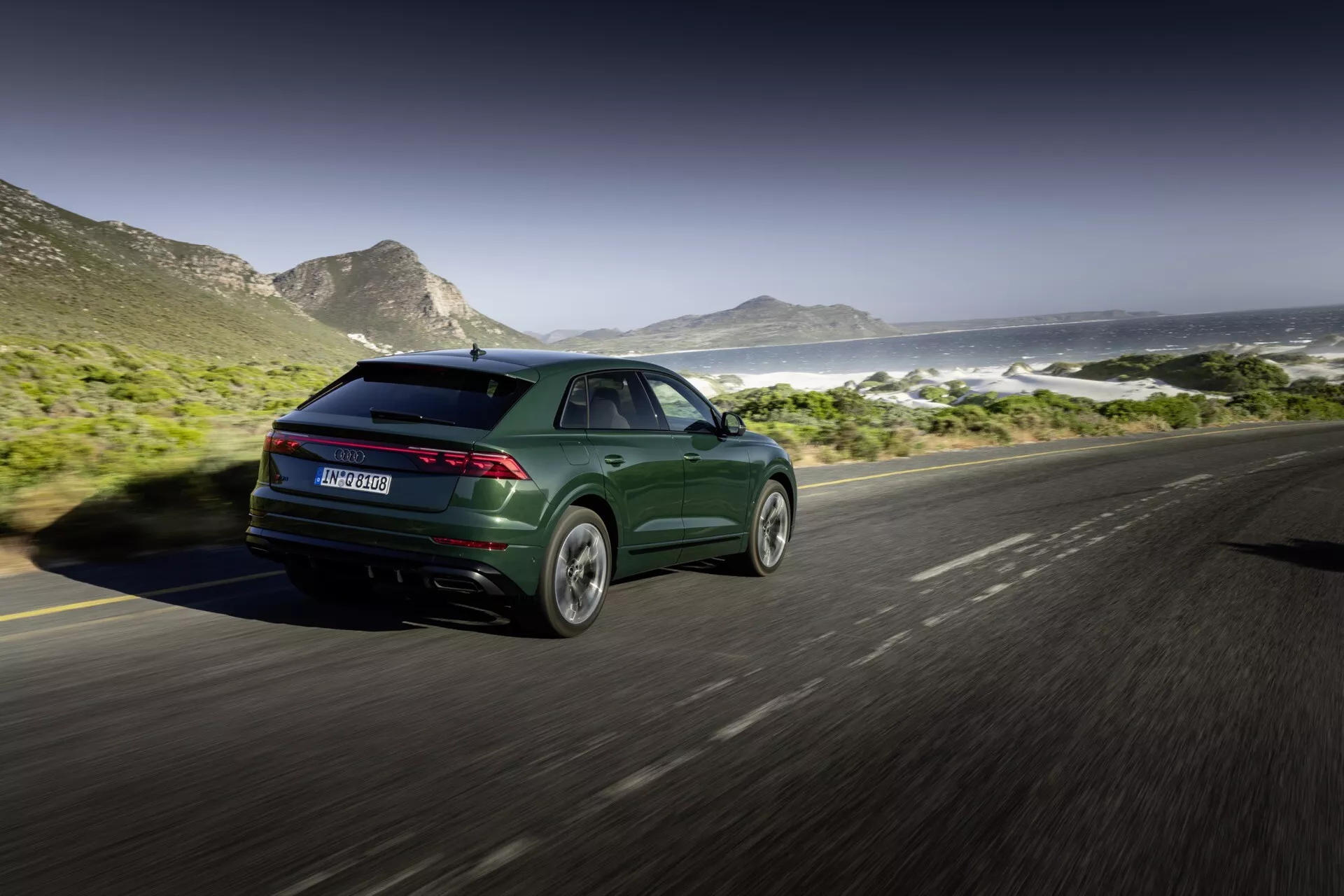 Audi Q8 2023 (4M-Facelift) v metalickom tmavo zelenej farbe, ukazuje svoj športový a moderný dizajn so štíhlymi zadnými svetlami a zliatinovými kolesami, zachytené z uhla späť tri štvrtiny na pobrežnej ceste s horami a oceánom v pozadí