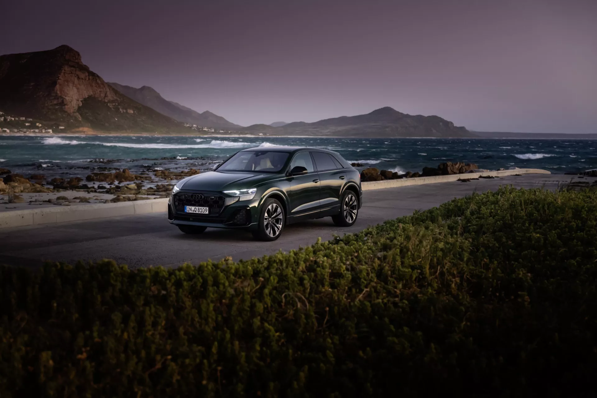 Audi Q8 2023 (4M-Facelift) tmavo sfarbený hatchback na malebnom pobrežnom ceste pri západe slnka, predstavujúci jeho LED svetlá, aerodynamický profil a zliatinové kolesá