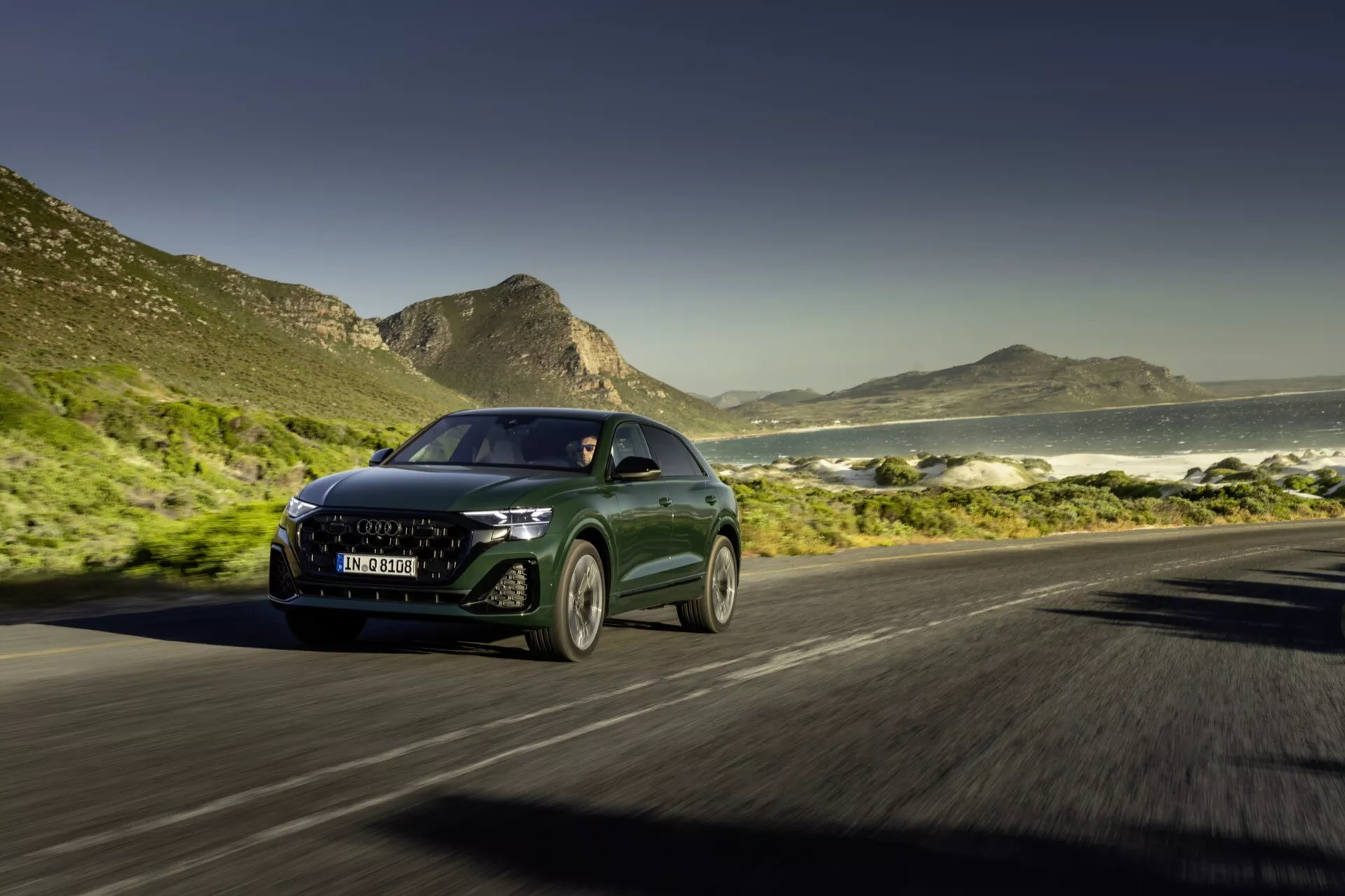 Audi Q8 4M Facelift 2023 idúce po pobrežnej ceste s dynamickým pohľadom spredu a z boku, ktorý ukazuje LED svetlomety, modernú mriežku a efekt rozmazania pohybu na pozadí oceánu a hôr za slnečného dňa