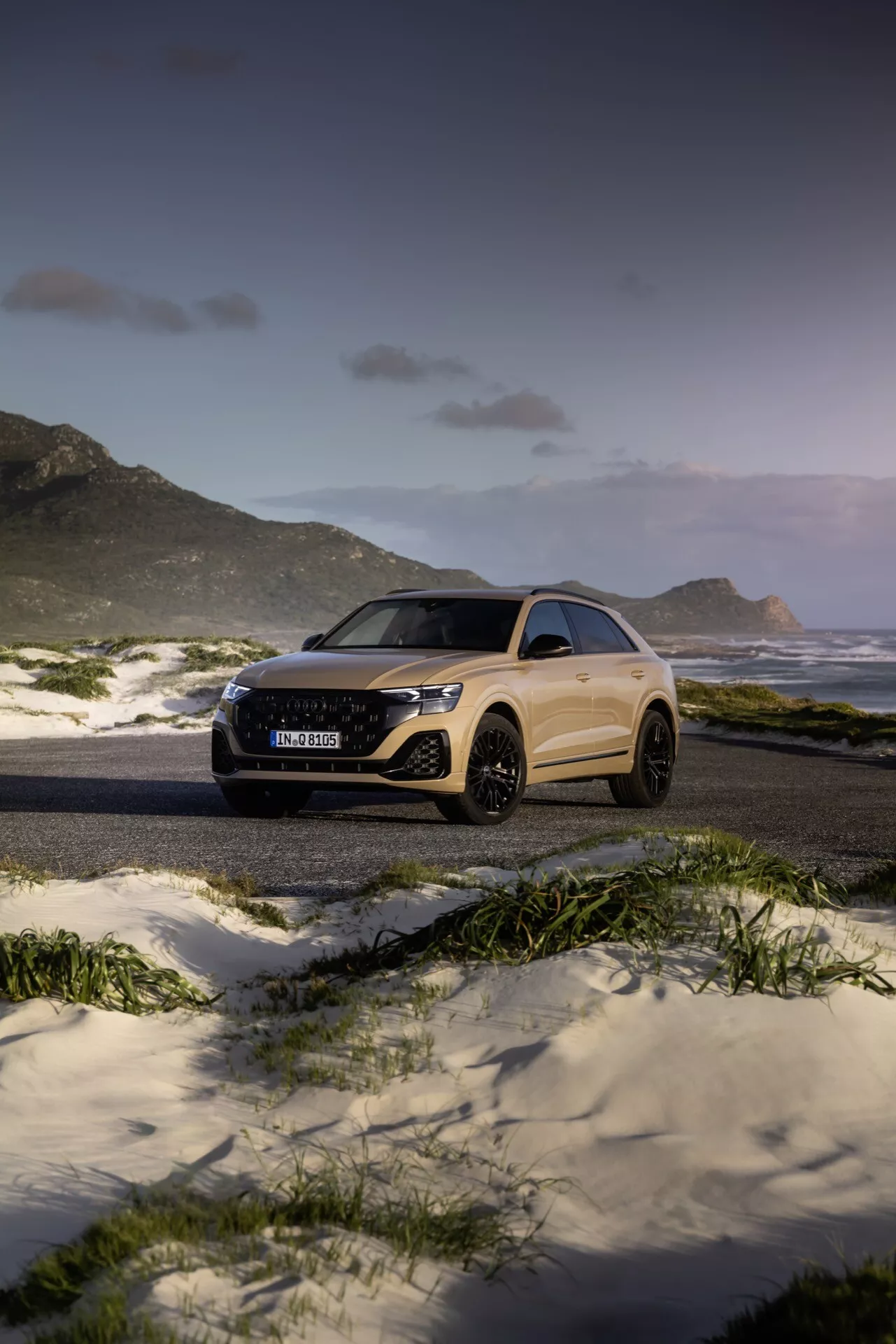 Audi Q8 2023 (4M-Facelift) v majestátnej plážovej a horskej krajine pri súmraku, predvádzajúce svoj elegantný dizajn a pokročilé funkcie, ako sú LED svetlomety a veľké zliatinové kolesá, z trojštvrtečného predného a bočného uhla