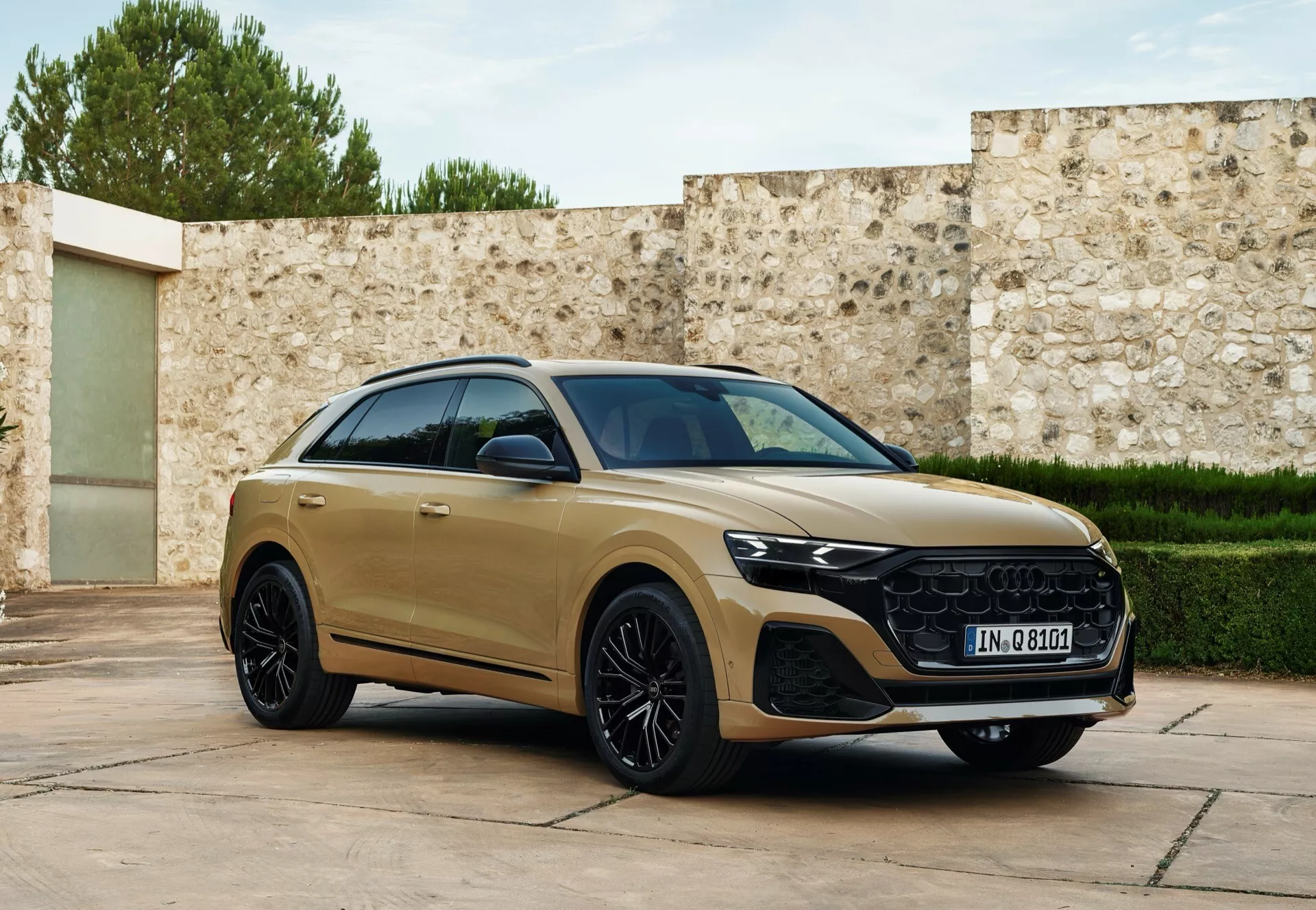 Trojštvrtečný predný pohľad na Audi Q8 2023 (4M-facelift) v zlatom odtieni, ukazujúci jeho aerodynamický bočný profil, charakteristickú mriežku, LED svetlomety a luxusné zliatinové kolesá, zaparkované s ľahkým otočením smerom k kamere na pozadí