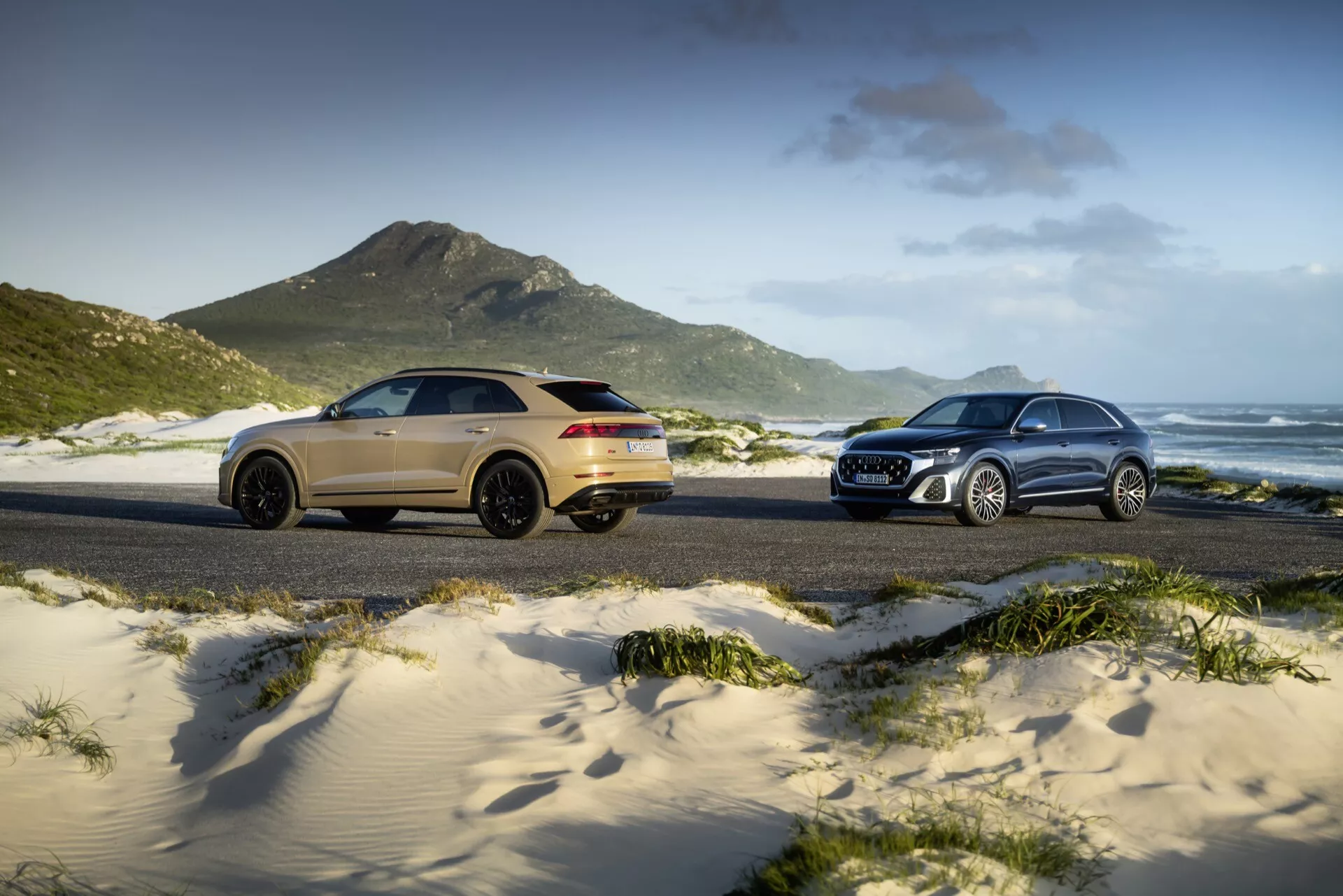 Modely Audi Q8 (4M-facelift) z roku 2023 predvádzané na piesočnatej pláži, so zlatým športovým SUV vľavo v trojštvrťovom zadnom výhľade a strieborným tradičným SUV vpravo v trojštvrťovom prednom výhľade, umiestnené proti trávnatým dunám, horám a čiastočne zamračenej oblohe