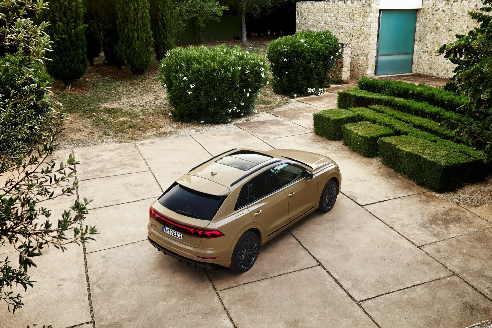 Sedan Audi Q8 2023 (4M-facelift) z vysokej zadnej trojštvrťovej perspektívy zobrazujúci LED zadné svetlá, panoramatické strešné okno a športové disky, zaparkovaný s malebným pozadím živých plotov a kamenné budovy