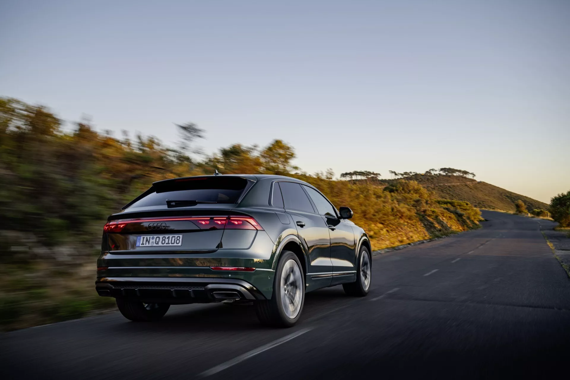 Audi Q8 2023 (4M-Facelift) v pohybe, zachytené z uhla tri štvrtiny zozadu, ktoré prezentuje moderné návrhové prvky vozidla vrátane elegantných, cez celú šírku rozťahujúcich sa zadných svetiel, dvojitých výfukových potrubí, zadného difúzora a integrovaného spoilera na pozadí prírodnej scenerie na otvorenej ceste pri západe alebo východe slnka, čo zdôrazňuje jeho orientáciu na výkon a pokročilú estetiku.