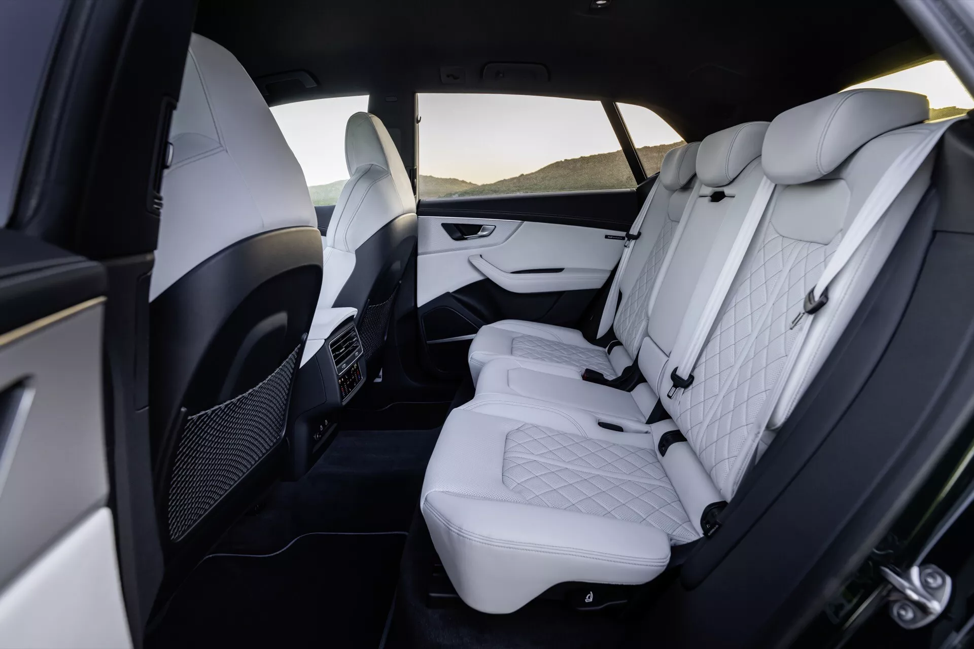 Pohľad na luxusný interiér zadného priestoru Audi Q8 2023 (4M Facelift), predstavujúci prémiovú bielu koženku s prešívaným diamantovým vzorom, nastaviteľné opierky hlavy, dvojfarebné dverové panely s chrómovými akcentmi, sklopnú lakťovú opierku, ventilácie klímy pre zadných pasažierov a priestranný priestor pre nohy.