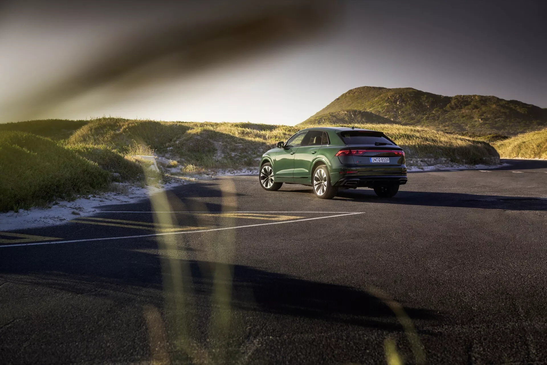 Zadný trojštvrťový pohľad na Audi Q8 (4M-Facelift) z roku 2023 na asfaltovej parkovacej ploche pri západe slnka, zvýrazňujúci jeho postoj, dizajnové čiary, LED zadné svetlá, zliatinové kolesá a leštený zelený povrch s prírodnou krajinou na pozadí.
