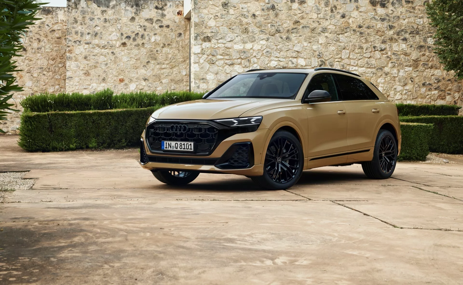 Luxusný crossover SUV Audi Q8 2023 v pohľade z troch štvrtín spredu zaparkovaný na dlažbe s pozadím starej kamennej steny, predstavujúci elegantné LED svetlomety, výraznú šesťuholníkovú mriežku, sochársky tvarované línie kapoty, veľké zliatinové kolesá a možný panoramatický strešný otvor.