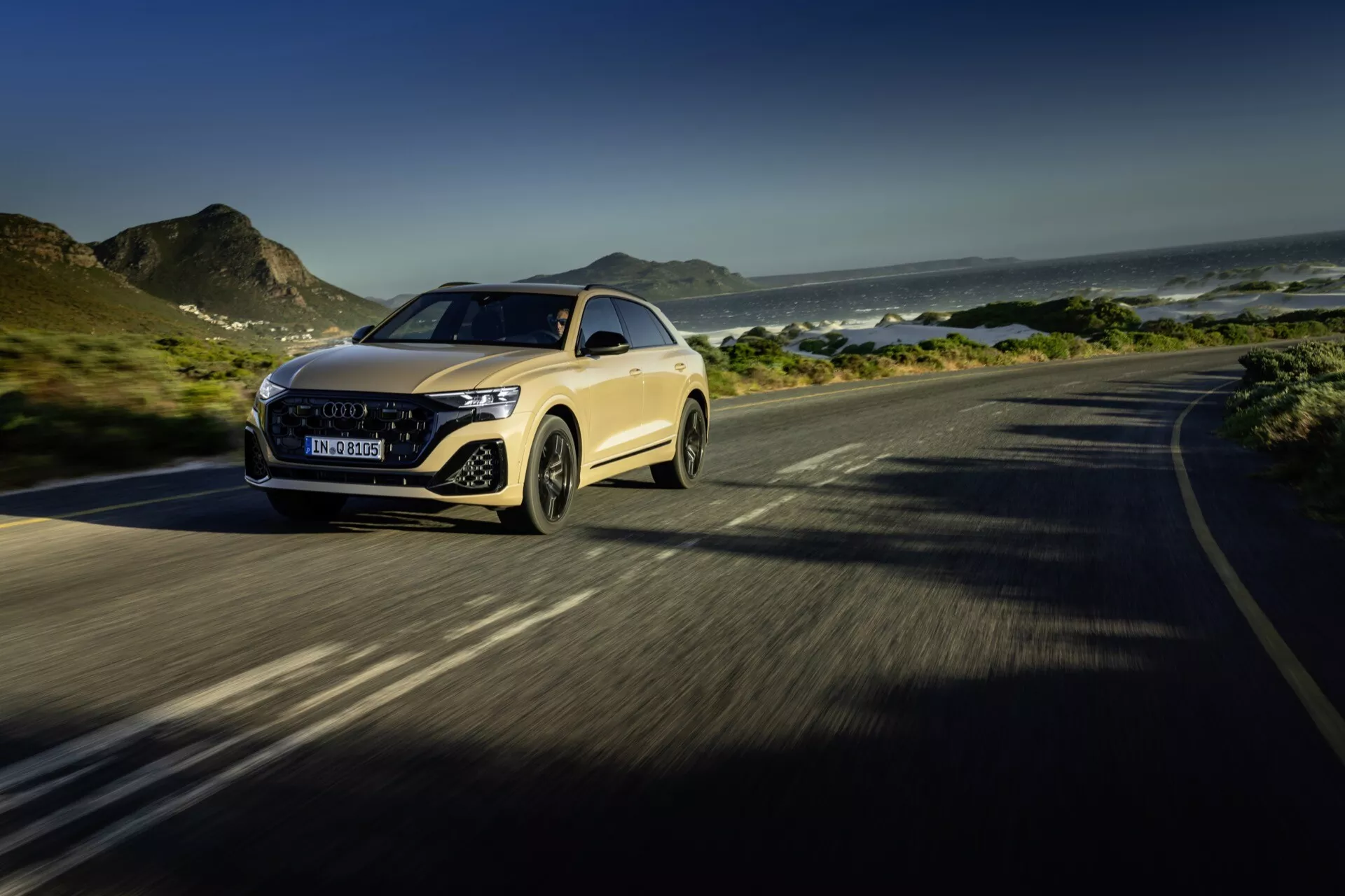 Audi Q8 4M facelift 2023 v pohybu na pobrežnej ceste, kde vynikajú pokročilé LED svetlomety luxusného auta, výrazná mriežka, aerodynamická kapota a dynamické ovládanie, nastavené proti pozadí hôr a oceánu so zameraním na dizajnové a technologické funkcie.