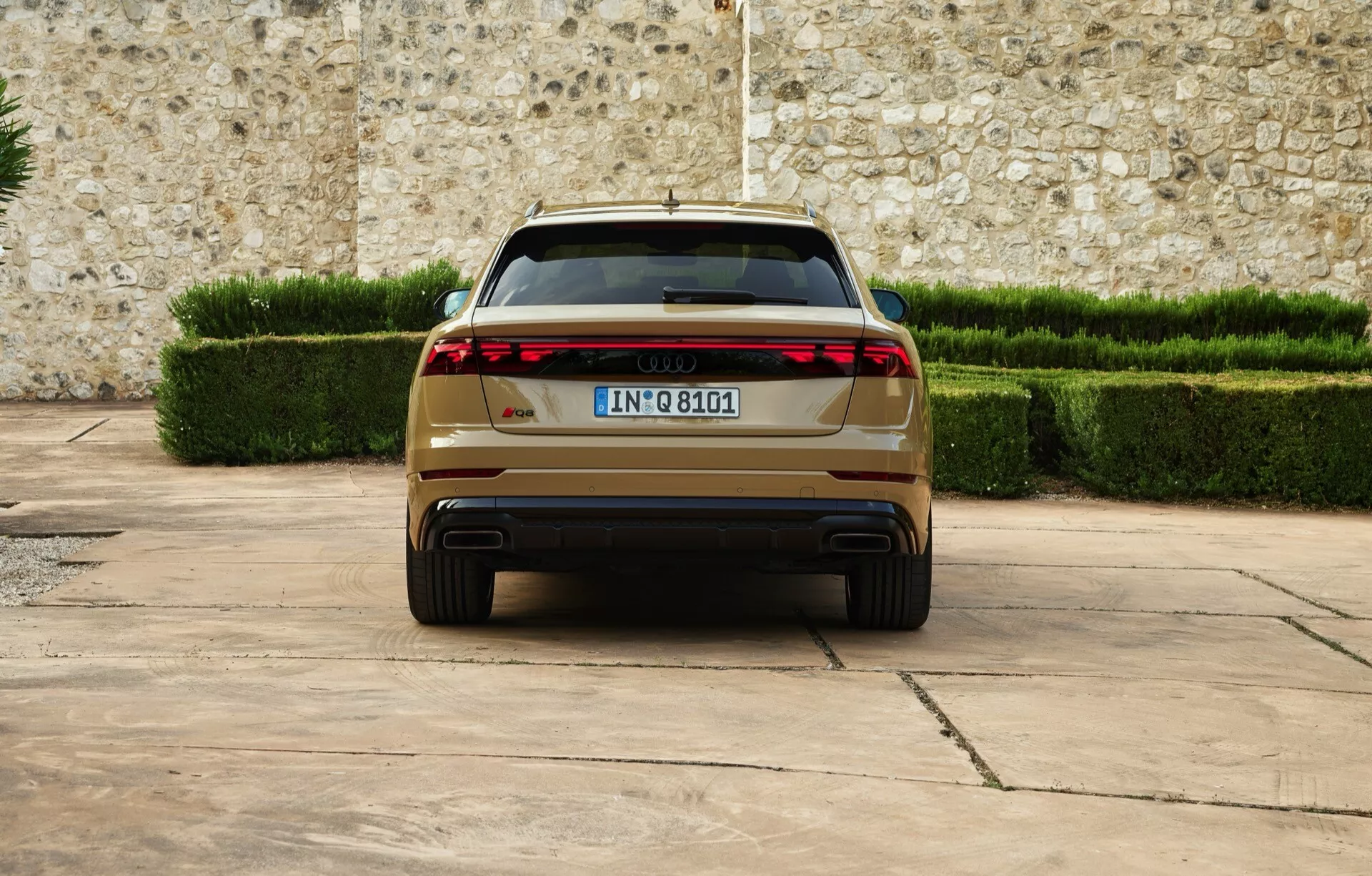Audi Q8 2023 (4M-facelift) ukazujúce zadný pohľad s modernými LED zadnými svetlami, dvojitými výfukmi, európskou evidenčnou značkou, zaparkované na spevnenej ploche proti pozadiu kamenného múru a upravených zelených živých plotov, čo zdôrazňuje luxus a pokročilé dizajnové funkcie