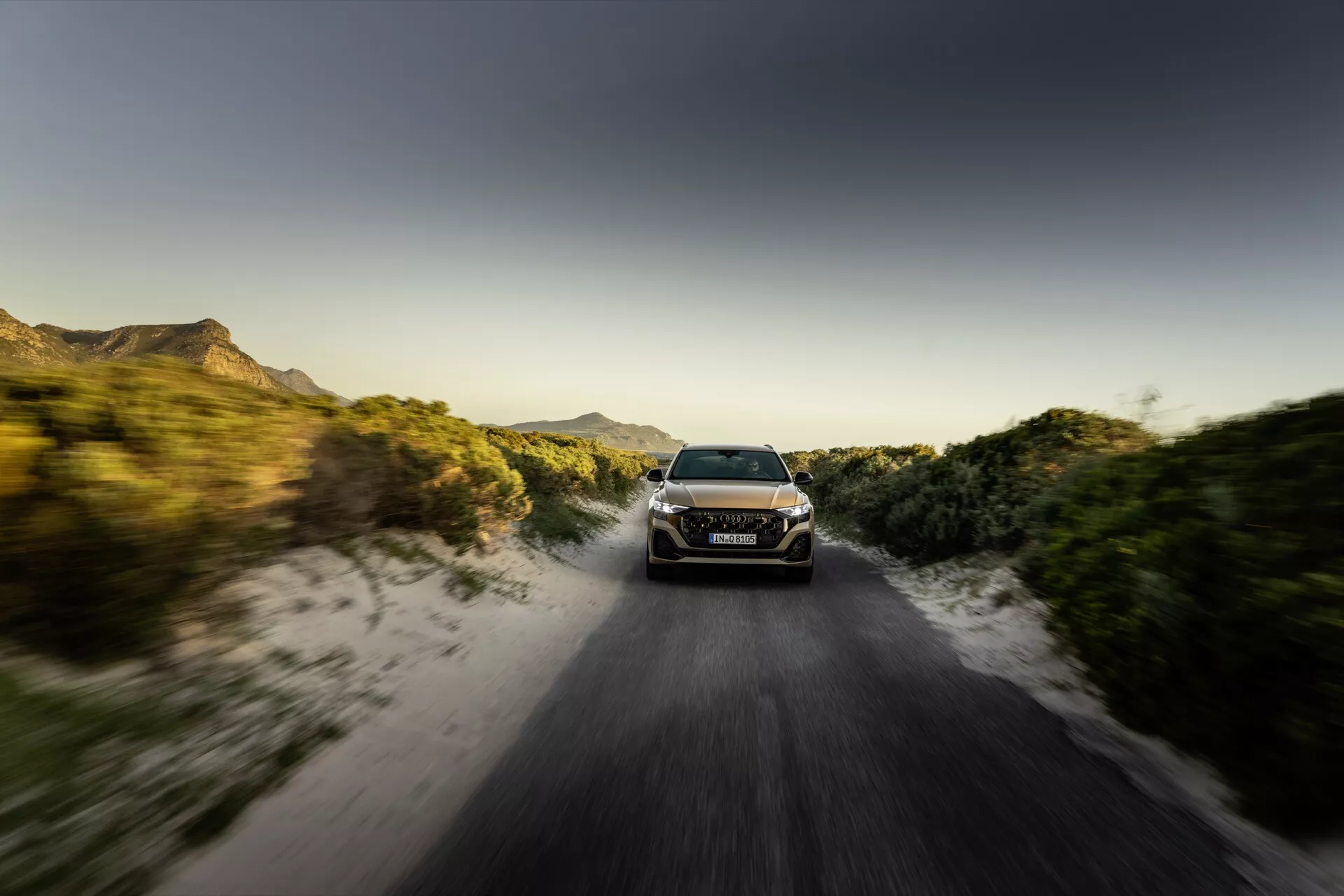 Audi Q8 2023 (4M-Facelift) v dynamickom pohybe na nezpevnené ceste pri východe alebo západe slnka s efektom rozmazania pohybu, ukazujúce jeho LED svetlomety, elegantný aerodynamický dizajn a schopnosť terénnej jazdy uprostred vidieckeho prostredia