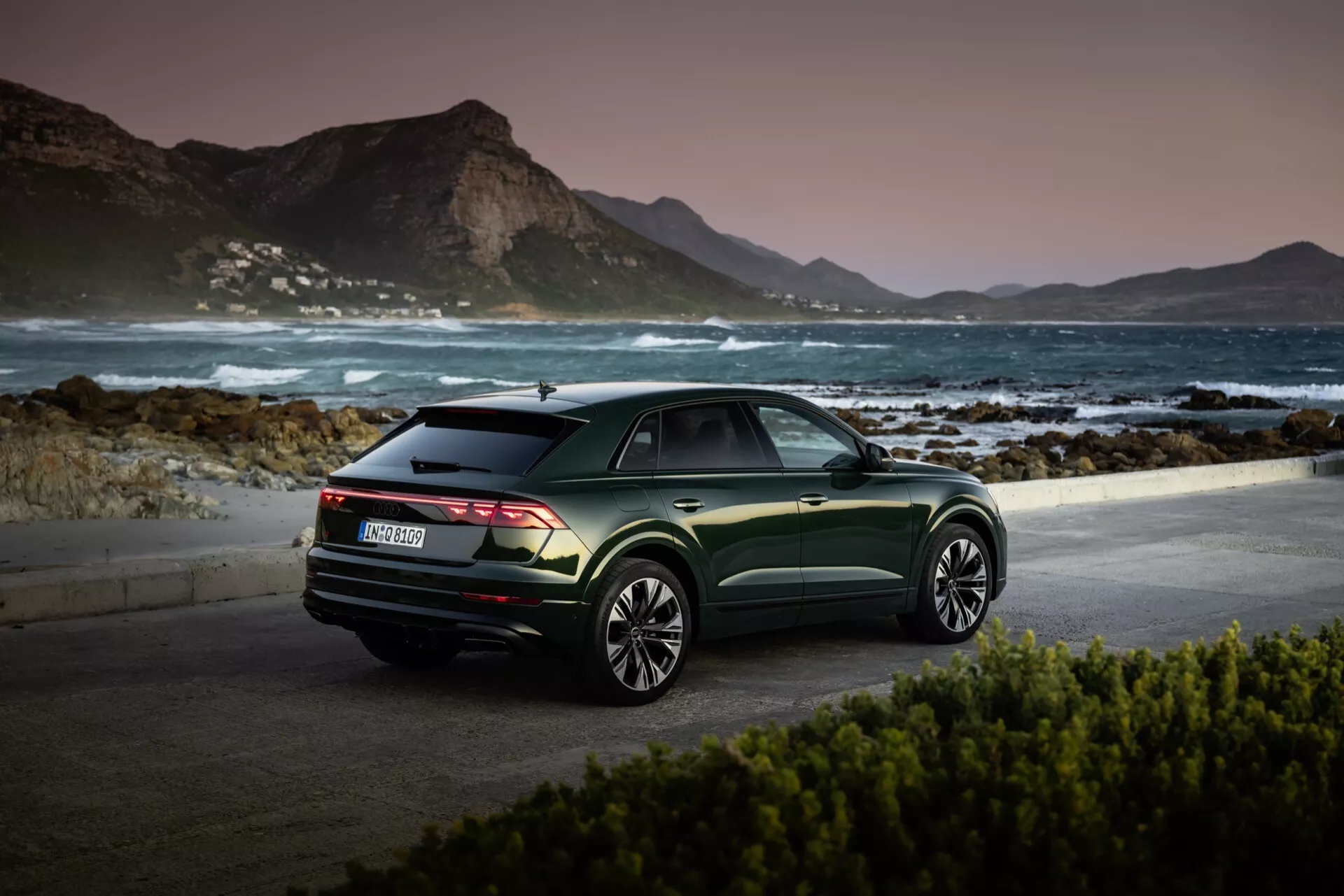 Audi Q8 2023 (4M-Facelift) zaparkované v malebnom prímorskom prostredí na úsvite alebo súmraku, predvádzajúce pohľad z troch štvrtín zadnej časti, tmavo zelené metalízovanej povrchovej úpravy a viacerých lúčových zliatinových kolies proti pokojnému horskému pozadiu
