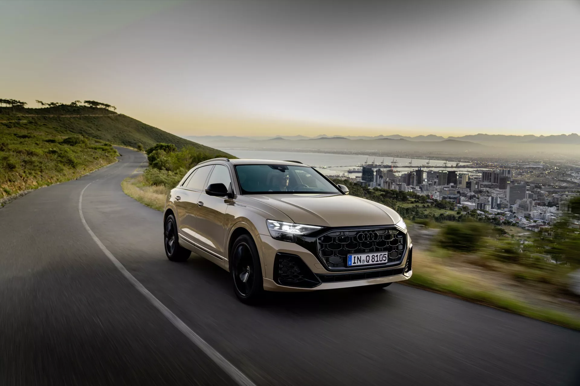Luxusné crossover SUV Audi Q8 2023 (4M-Facelift) v pohybe na zákrutej ceste, predstavuje športový dizajn, LED svetlomety, veľké zliatinové kolesá a panoramatickú strechu, proti pozadiu mesta a hôr v mäkkom rannom alebo neskorom popoludňajšom svetle.