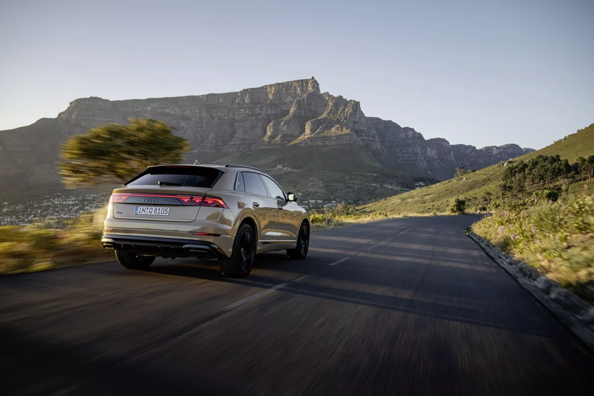 Model Audi Q8 4M facelift 2023 v dynamickom pohybe na zákrutej ceste, zdôrazňujúci jeho moderný dizajn SUV a pokročilé funkcie z pohľadu z troch štvrtí zozadu, s malebným horským pozadím pri západe slnka