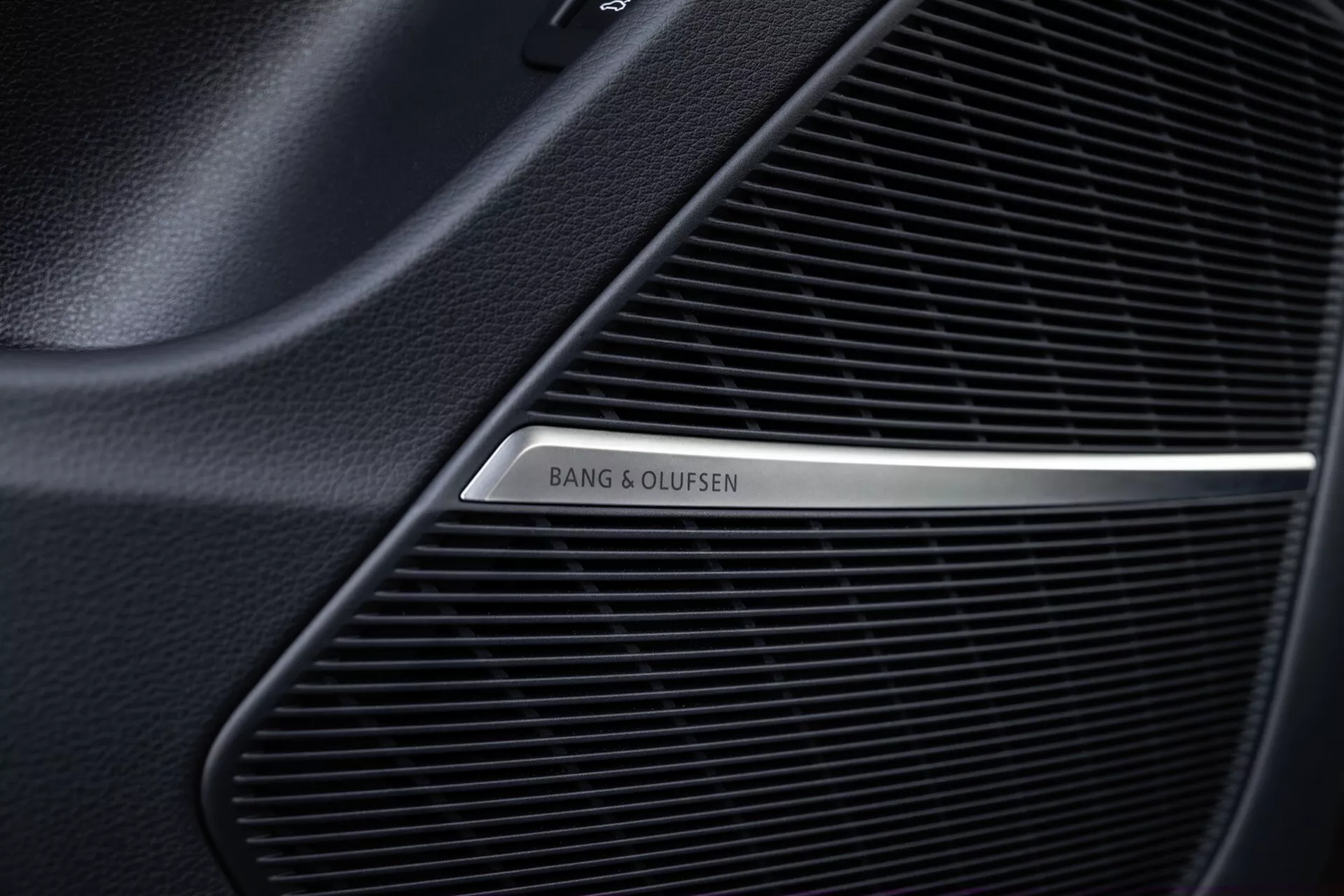 Záber zblízka na mriežku reproduktora Bang & Olufsen v interiéri Audi Q8 2023 (4M-Facelift), ukazujúci jemný mriežkový vzor prémiového zvukového systému a leštené kovové logo na čiernom pozadí.