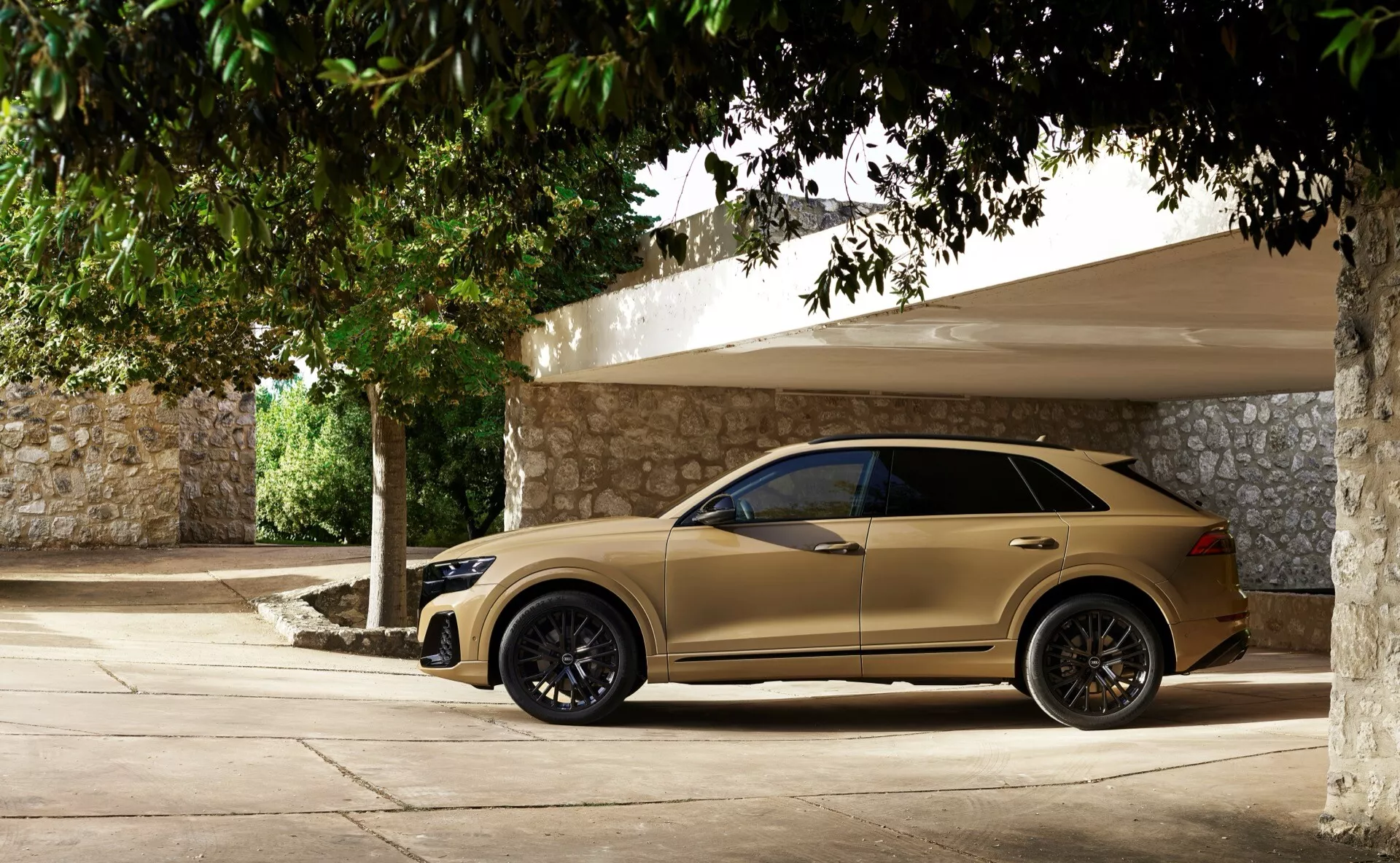 Audi Q8 2023 (4M-Facelift) v béžovej/zlatej farbe, zobrazuje celý bočný pohľad s tmavými zliatinovými kolesami, zrkadlami s integrovanými smerovými signálmi, tónovanými oknami, zapustenými kľučkami dverí a sklonenou strechou, zaparkované v tieni na slnečný deň