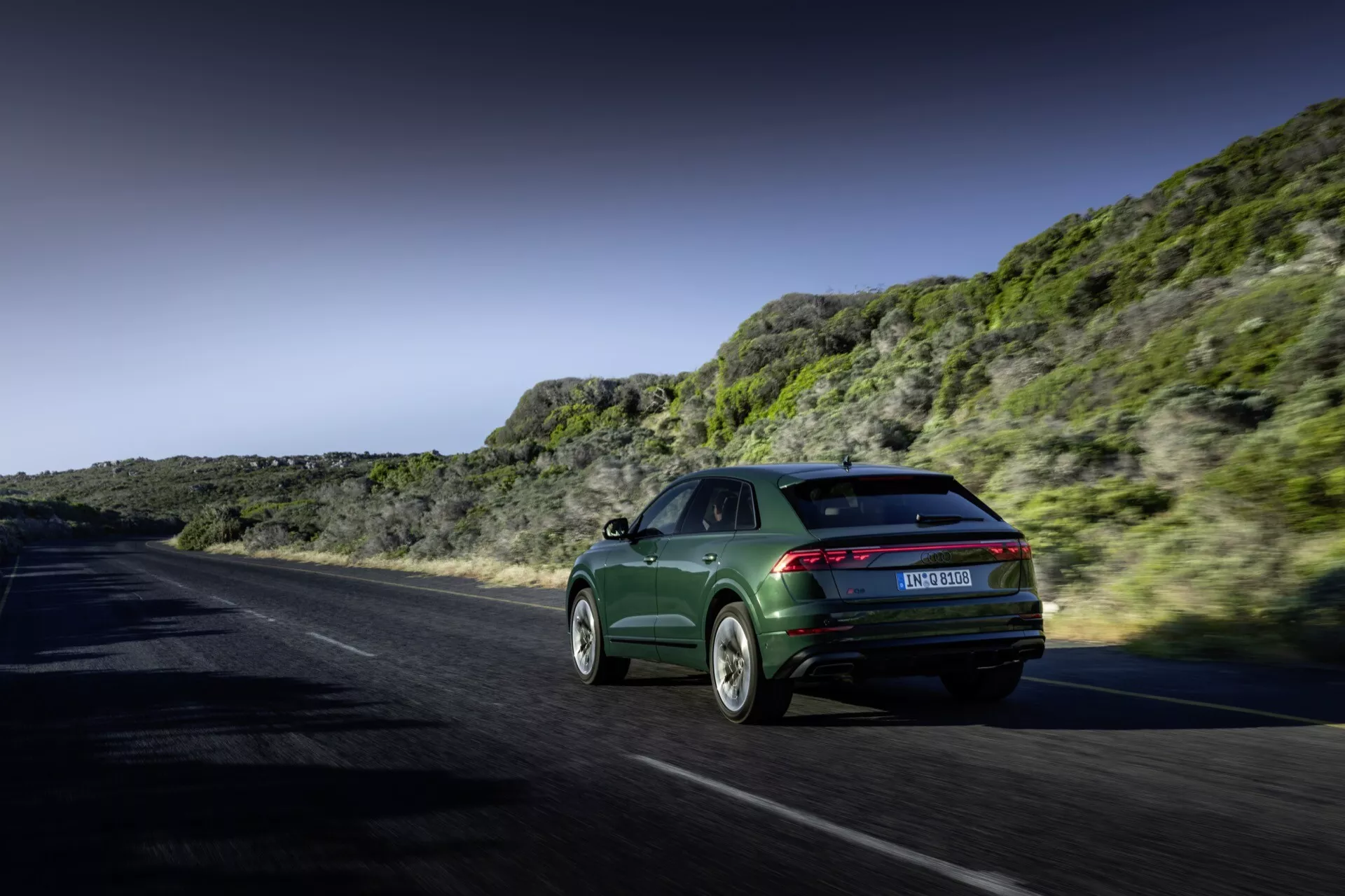 Audi Q8 2023 (4M-Facelift) v dynamickom pohybe na pobrežnej alebo horskej ceste, ukazujúci jeho štýl sportback, elegantné zadné svetlá a sofistikované zliatinové kolesá proti dramatickej krajine