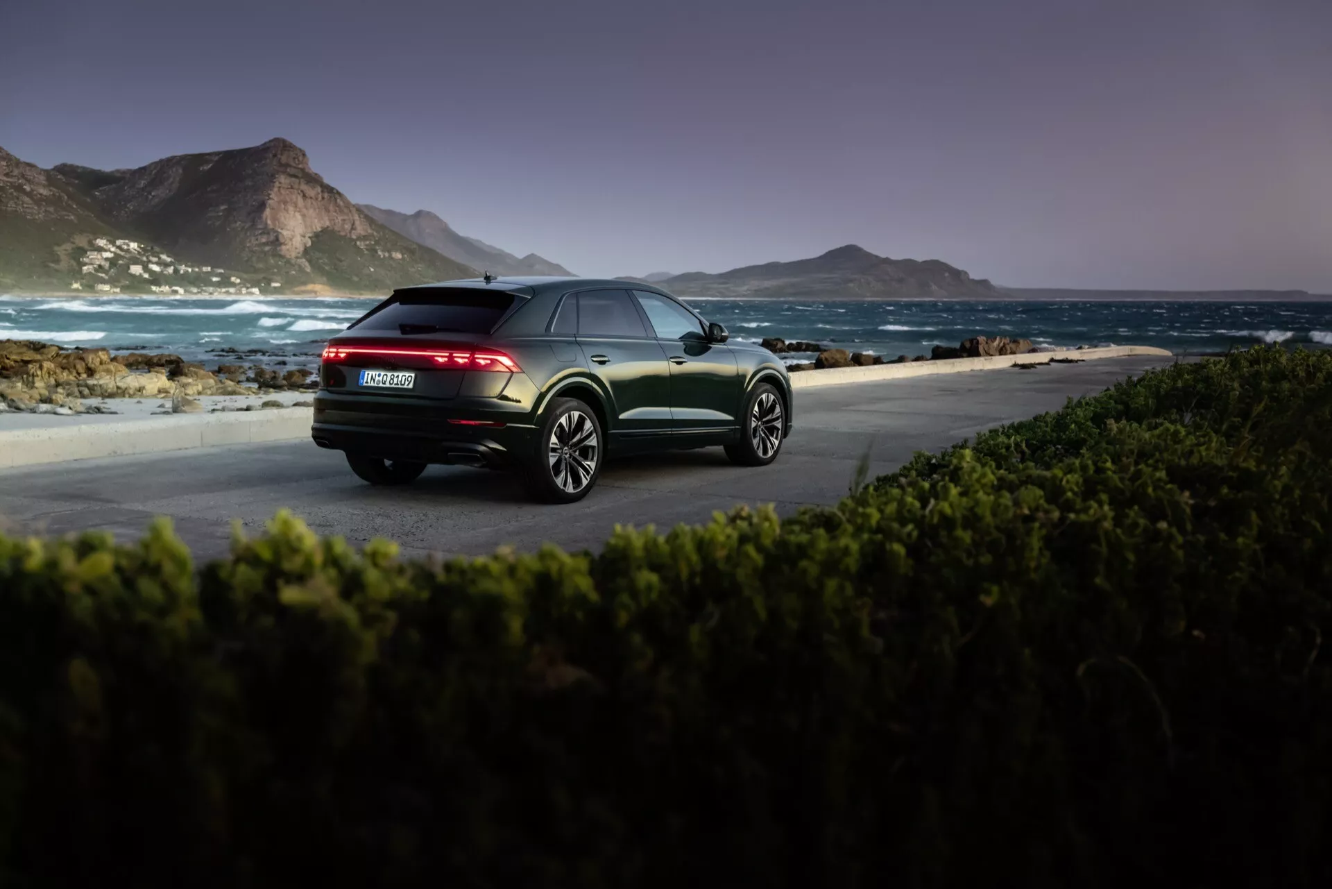 Audi Q8 4M-Facelift 2023 s metalickým povrchom zachytené na úsvite alebo súmraku v prírodnom pobrežnom prostredí, zdôrazňujúce zadný trojštvrtečný pohľad s LED zadnými svetlami, dvojitými výfukmi a zliatinovými kolesami s viacerými lúčmi.