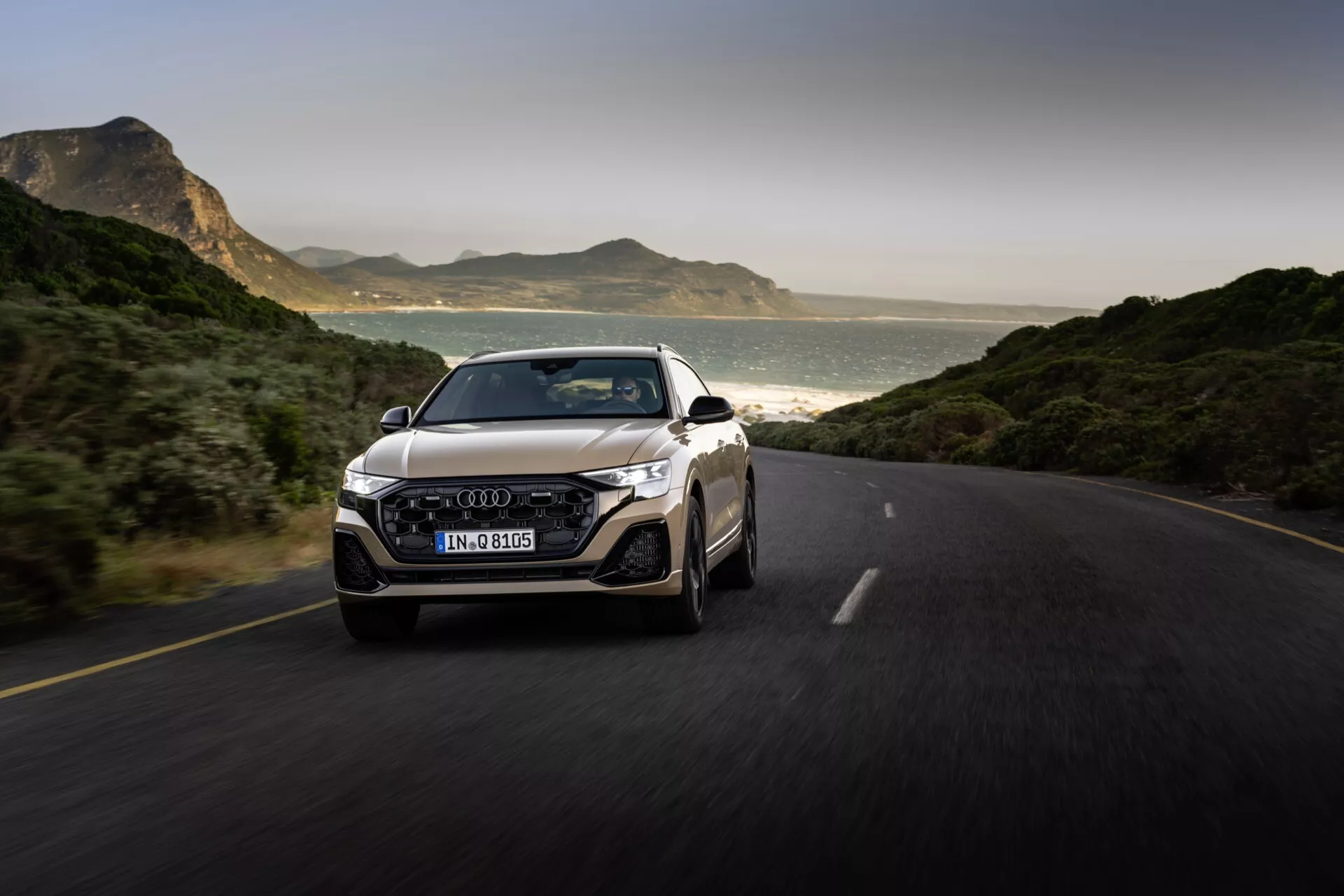 Audi Q8 2023 (4M-Facelift) zachytené v pohybe na zpevnenej ceste, prezentuje jeho dynamický dizajn z pohľadu troch štvrtín zpredu, s dôrazom na mriežku, LED svetlomety, nárazník a emblém na mriežke, so scenickým pozadím hôr a pobrežia pri prirodzenom osvetlení