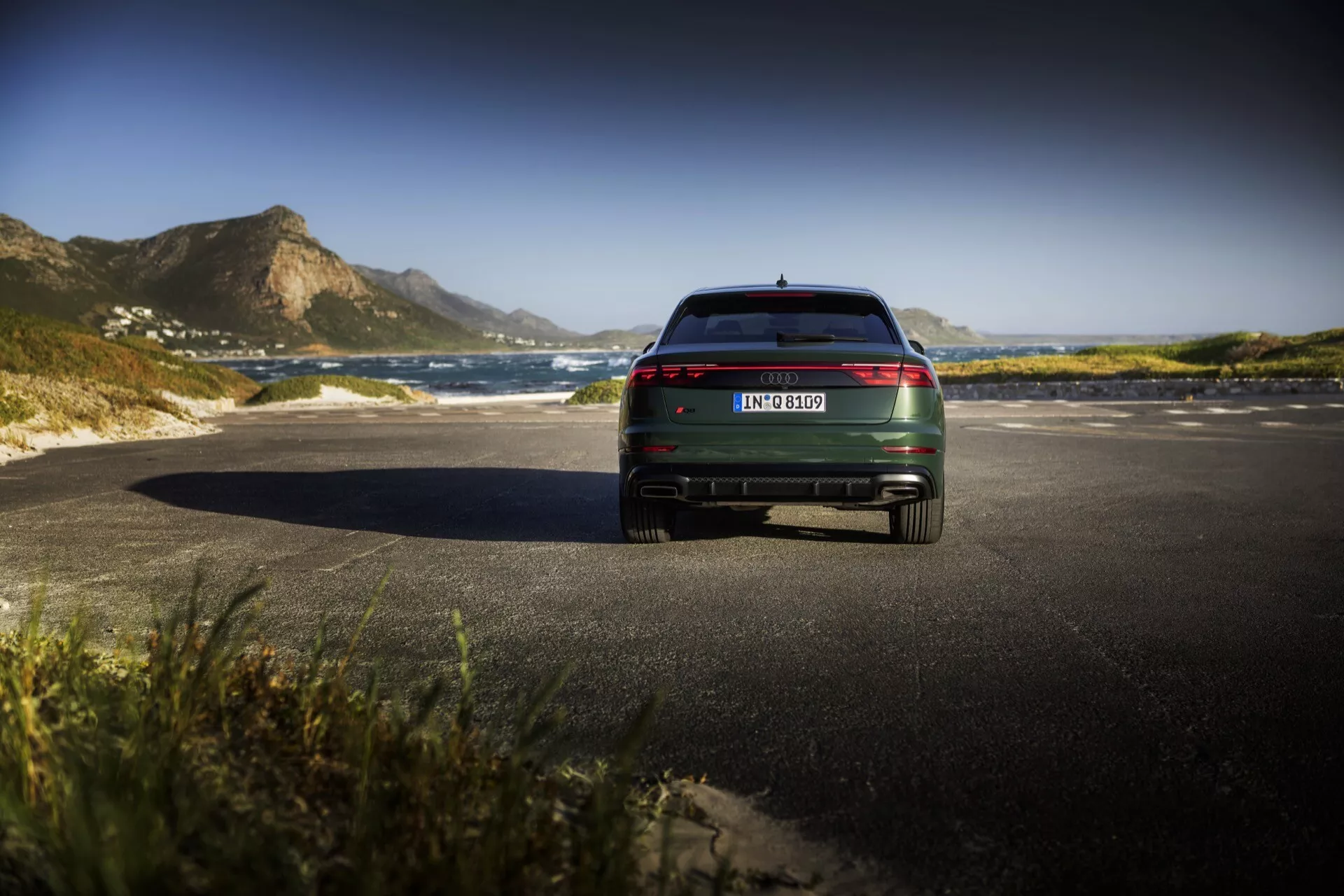 Zadný pohľad na model Audi Q8 4M Facelift 2023, s LED zadnými svetlami, chrómovými akcentmi a dvojitým výfukom na malebnej pobrežnej ceste