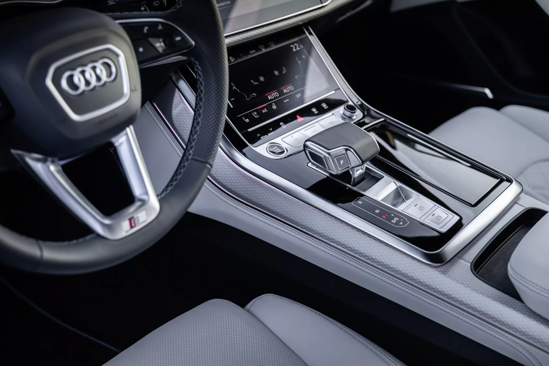Detailný pohľad na stredovú konzolu a časť volantu Audi Q8 2023 (4M-Facelift) z pohľadu spolujazdca, zvýrazňujúci tlačidlá na multifunkčnom volante, centrálny dotykový displej infotainmentu, oblasť regulácie klímy a modernú radicu páku vo vysokej kvalite interiérového prevedenia.