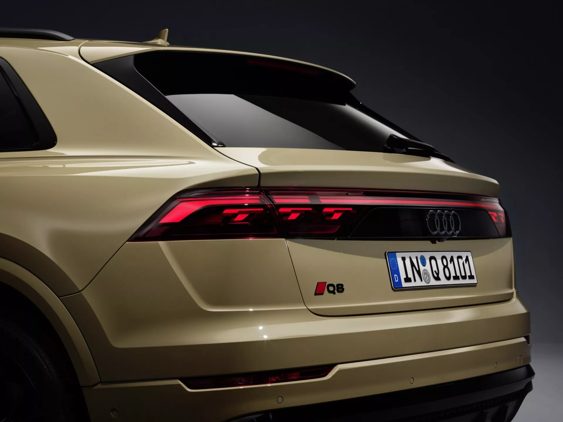Zadný trojštvrťový pohľad na Audi Q8 4M-Facelift rok 2023 s LED zadnými svetlami, elegantným zadným nárazníkom s integrovanými snímačmi, označením Audi, európskou evidenčnou značkou, zadným spoilérom, zlatavo-bežovou farbou laku a anténou v tvare žraločej plutvy na neutrálnom šedom pozadí.