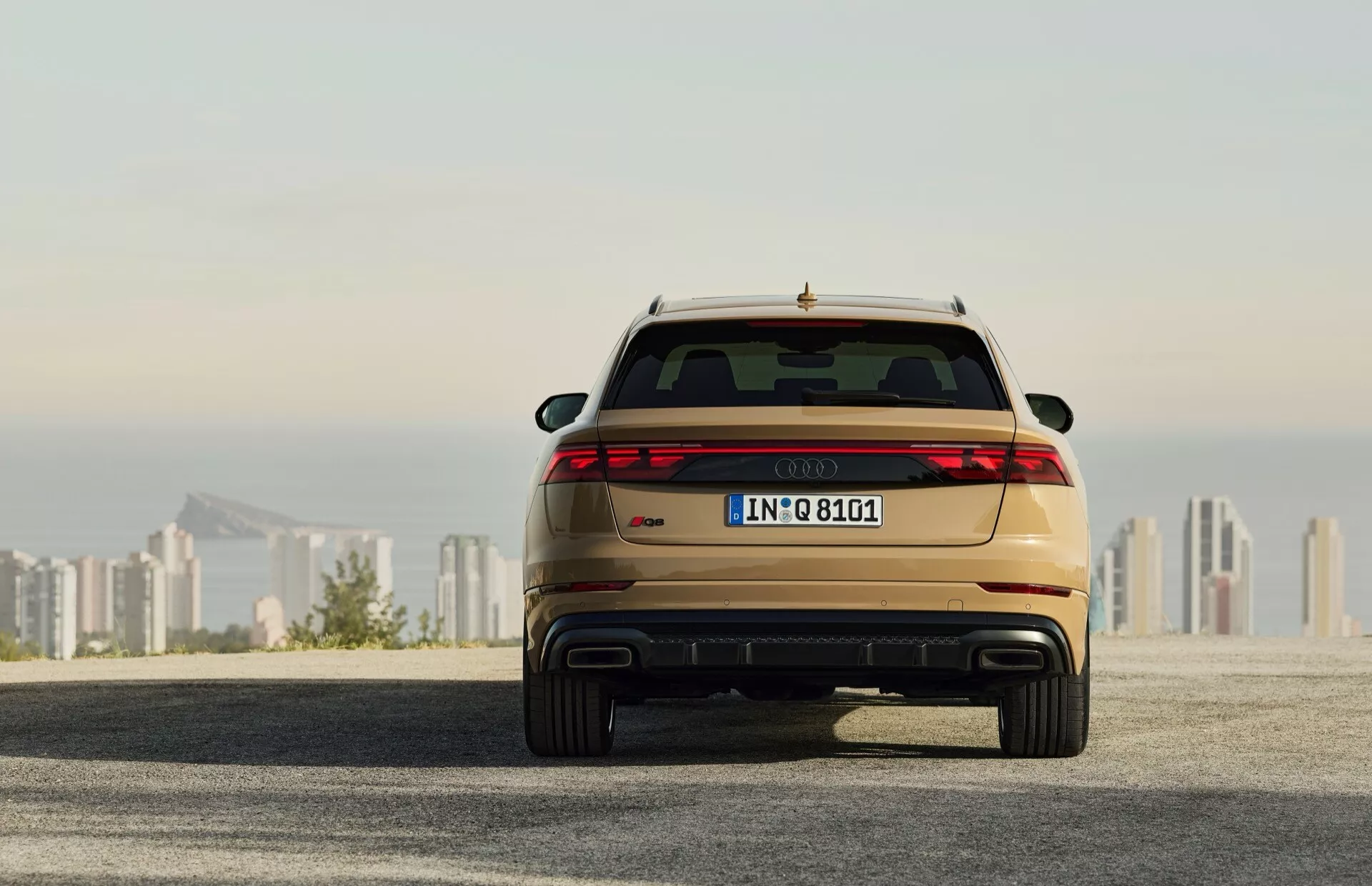 Zadný pohľad na Audi Q8 2023 (4M-Facelift) ukazujúci LED zadné svetlá, dvojitý výfuk a metalický lak, zaparkované na asfaltovom povrchu s panorámou mesta na pozadí počas východu/západu slnka