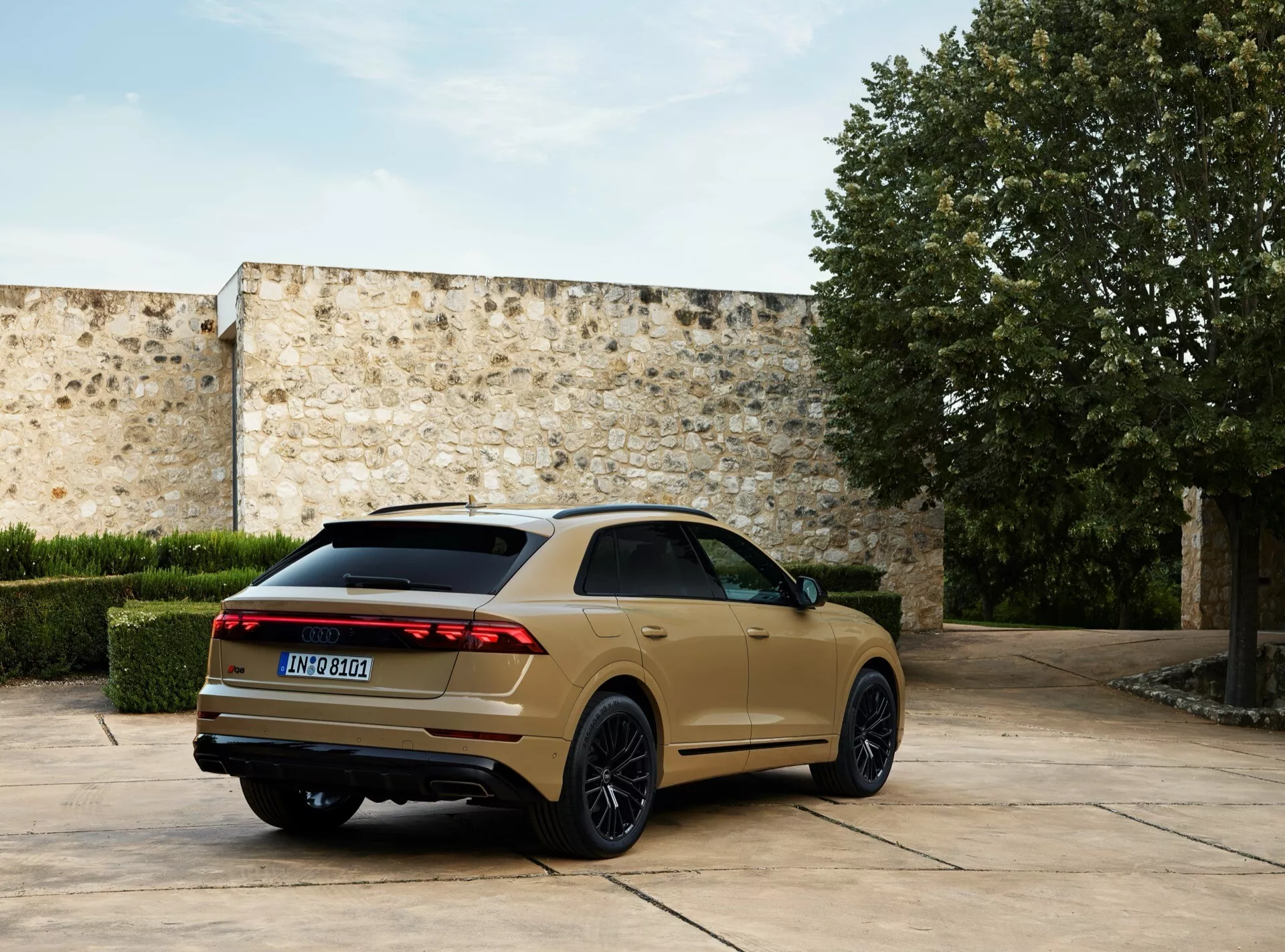 Model Audi Q8 4M facelift z roku 2023 zaparkovaný vonku, zachytený z uhla tri štvrtiny zozadu, ktorý zdôrazňuje jeho LED zadné svetlá, tmavé zliatinové kolesá s viacerými paprskami, sklonenú strechu a dvojité výfukové potrubia na pozadí kamenného nádvoria