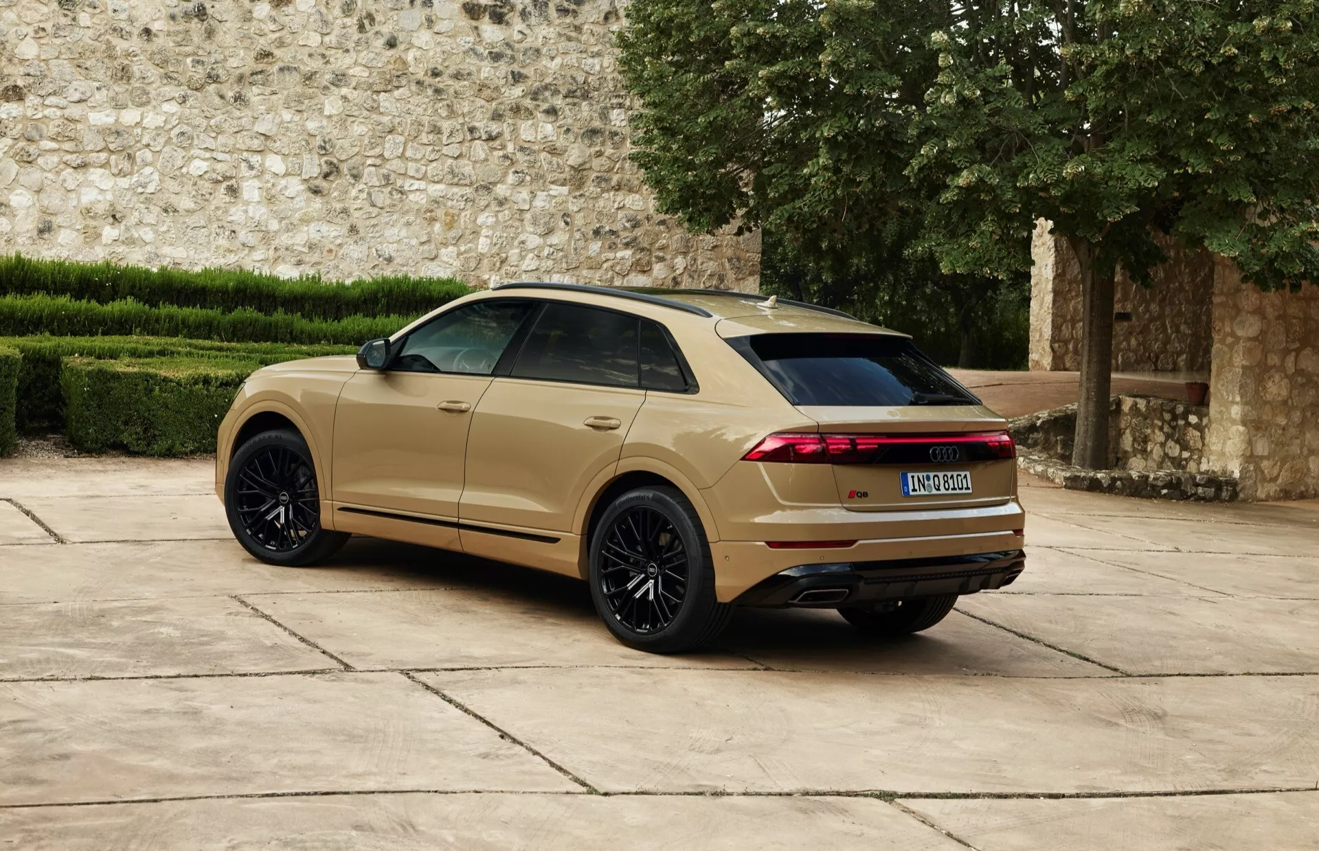 Audi Q8 2023 (4M-Facelift) zobrazené z trojštvrtečného zadného pohľadu ukazujúce veľké čierne viacramenné zliatinové kolesá, metalický zlatisto-bežový lak, predĺžené horizontálne LED zadné svetlá, zadný spoiler a dvojitý výfukový systém na pozadí historického kameňa múr
