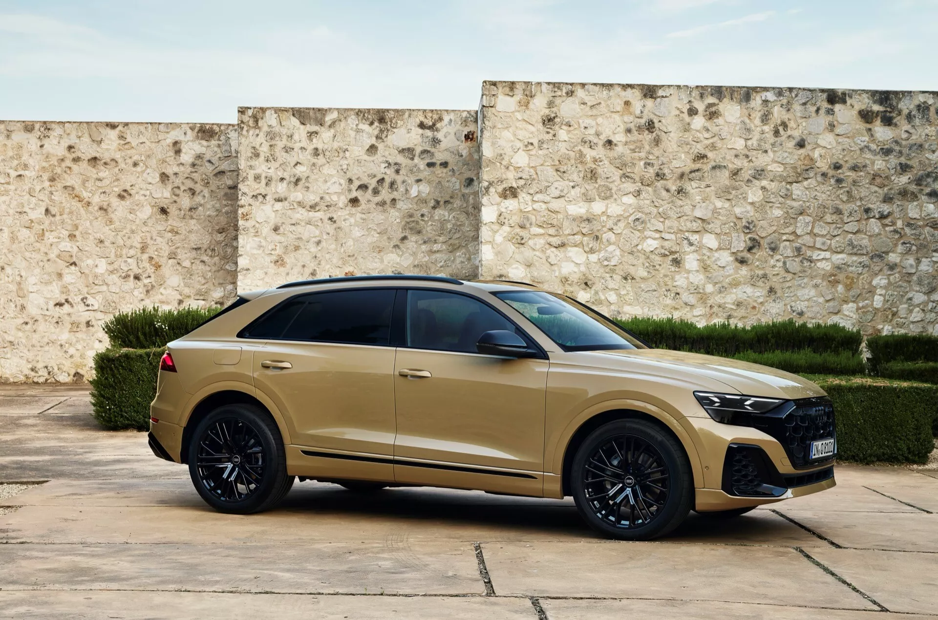 Trojštvrťový predný pohľad na moderné Audi Q8 2023 (4M-Facelift) v matnej zlatej alebo béžovej farbe, s veľkou mriežkou s medovcom, LED svetlometmi, športovým predným nárazníkom, čiernymi zliatinovými kolesami, pneumatikami s nízkym profilom, zapustenými kľučkami dverí, tmavo tónovanými oknami, sklápacou strešnou líniou v štýle kupé, svalnatou bočnou líniou s uhlovými zadnými svetlami a strešným spojlerom, pred textúrovanou stenou naznačujúcou historické alebo súčasné prostredie.
