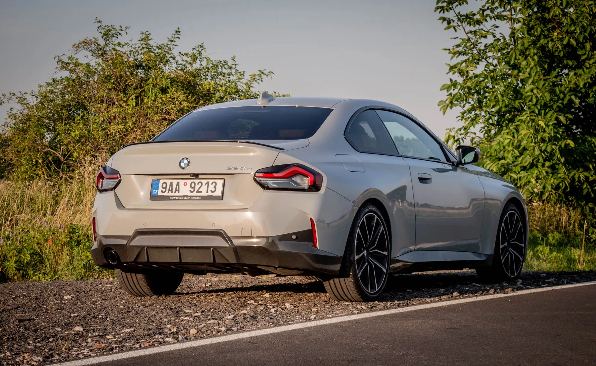 Pohľad zozadu na športové vozidlo BMW 2er-reihe 2022 (2-serie coupe) zachytený z nízkeho uhla. Auto, zaparkované na okraji cesty v vidieckom prostredí s prírodnou zeleňou, je viditeľné pri mäkkom osvetlení. Výrazné odznaky identifikujú značku a model, zobrazujúc zadnú časť vrátane veka kufra, zadného nárazníka a obchádzajúcich zadných svetiel. Dvojité výfukové potrubia naznačujú výkonný motor. Malý integrovaný zadný spojler a veľké zliatinové kolesá s tenkými lúčmi naznačujú aerodynamický štýl a výkon. Kovová farba a hladko klesajúca strešná línia vozidla zvyšujú jeho prémiový vzhľad. Možné moderné funkcie viditeľné zahŕňajú LED zadné svetlá, parkovacie senzory a kameru na cúvanie.