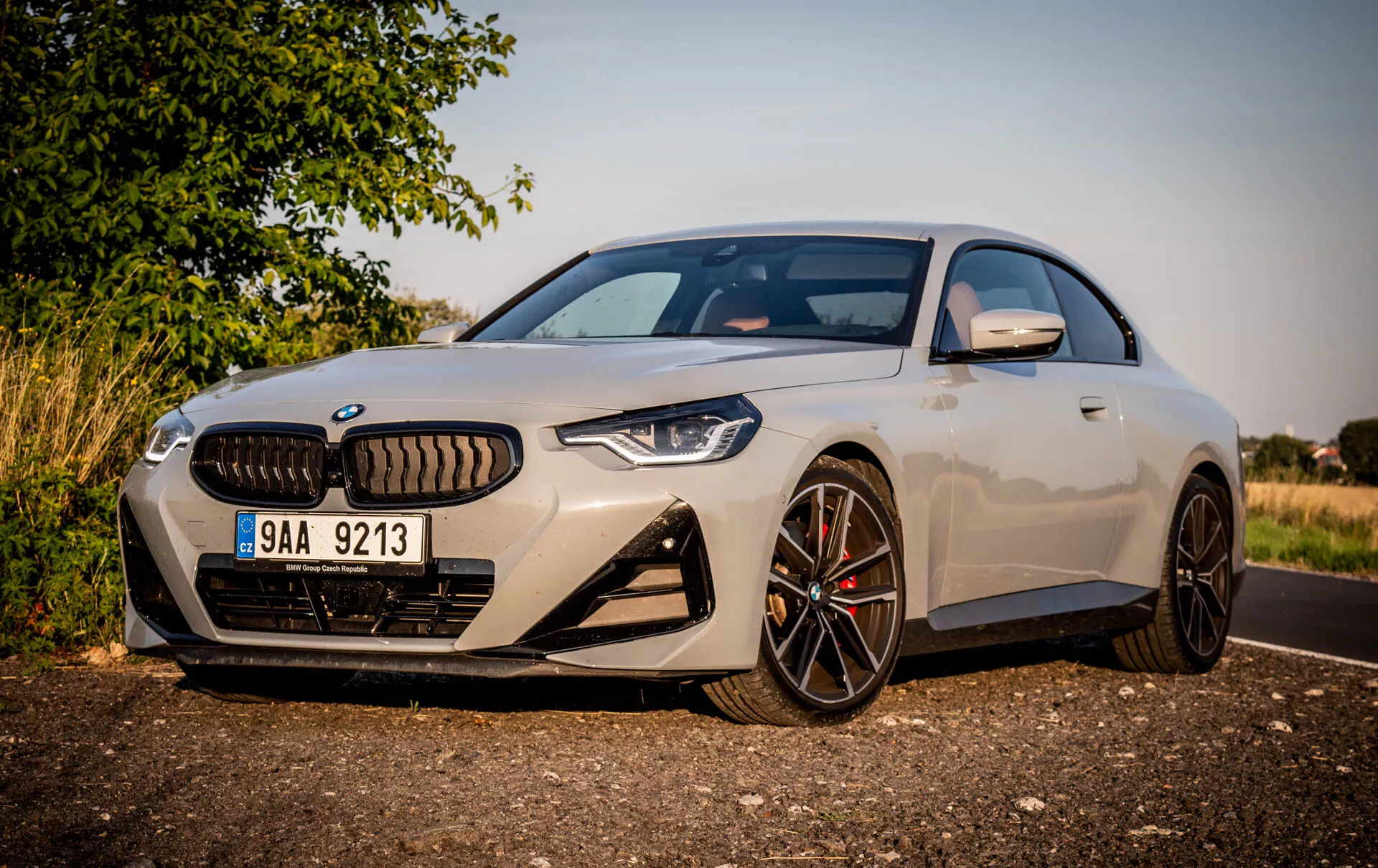 Obrázok moderného športového kupé BMW radu 2 z roku 2022. Auto je zaparkované na kraji cesty počas dňa, zobrazené pod šikmým uhlom z ľavej prednej strany, čím vyniká strana vodiča. Funkcie zahŕňajú elegantný aerodynamický dizajn, veľké viacramenné kolesá z ľahkej zliatiny s tmavou povrchovou úpravou, kontrastné brzdové strmene, charakteristickú mriežku chladiča obklopenú úzkymi agresívnymi LED svetlometmi, prednú kameru nad poznávacou značkou, veľké vstupy do predného nárazníka, svetlý metalický lak, spätné zrkadlá s integrovanými smerovkami a čiastočný pohľad na panoramatickú strechu. Auto je umiestnené blízko kamery s vidieckym alebo prímestským pozadím, teplým osvetlením a tieňmi, ktoré naznačujú nízke slnko buď ráno alebo neskoro popoludní.