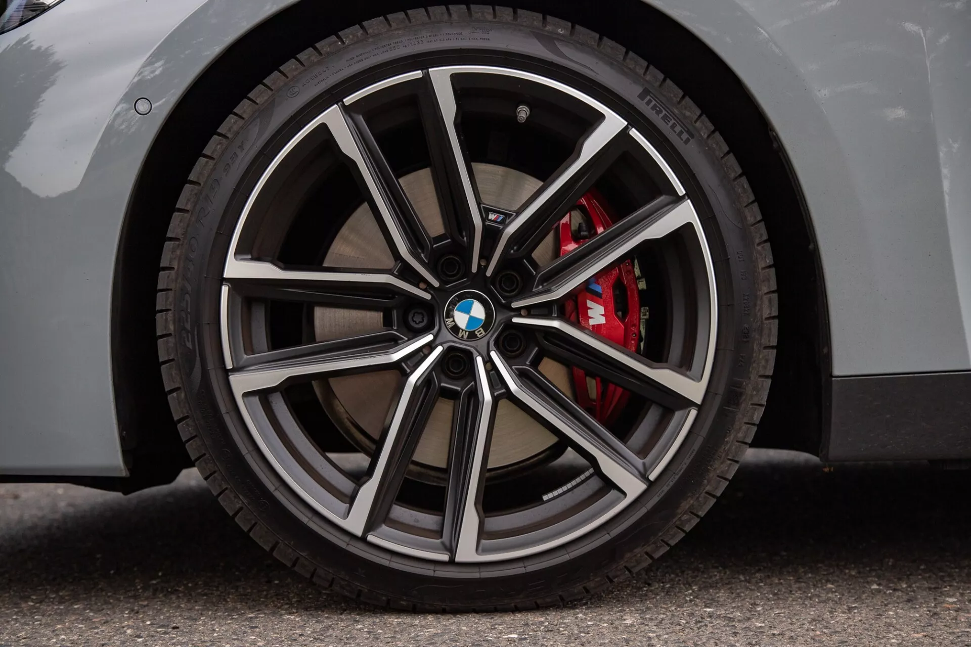 Detailný záber na koleso BMW 2 Series Coupé s viacramenným diskovým dizajnom s leštenými a tmavšími kovovými povrchmi, logom BMW na kryte, pneumatikou s nízkym profilom a červeným brzdovým strmeňom. Svetlá karoséria na asfaltovej ploche zdôrazňuje luxusné a športové aspekty.