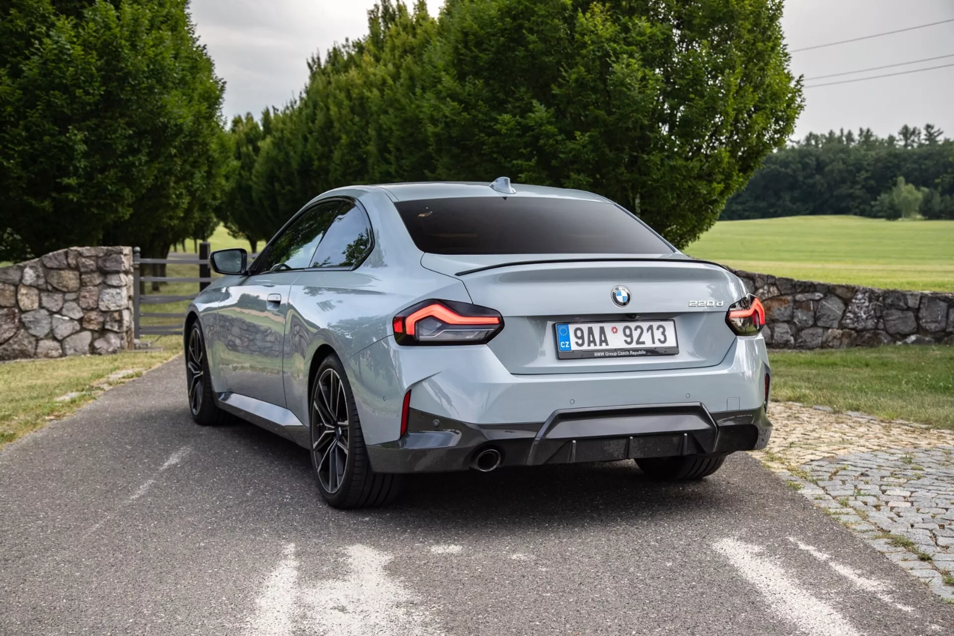 Obrázok BMW 2er-Reihe 2022 2-Series Coupe zachyteného zo zadného trojštvrťového uhla. Auto, umiestnené takmer diagonálne, ukazuje výrazné LED zadné svetlá, dva výfukové otvory, viacramenné zliatinové kolesá a lesklý metalický lak. Zadný dizajn zahŕňa emblém výrobcu v strede a odznak modelu/motorovej varianty na veku kufra. Vonkajšie nastavenie zahŕňa dláždenú cestu alebo príjazdovú cestu, trávnatú plochu, stromy a čiastočne viditeľnú kamennú stenu.