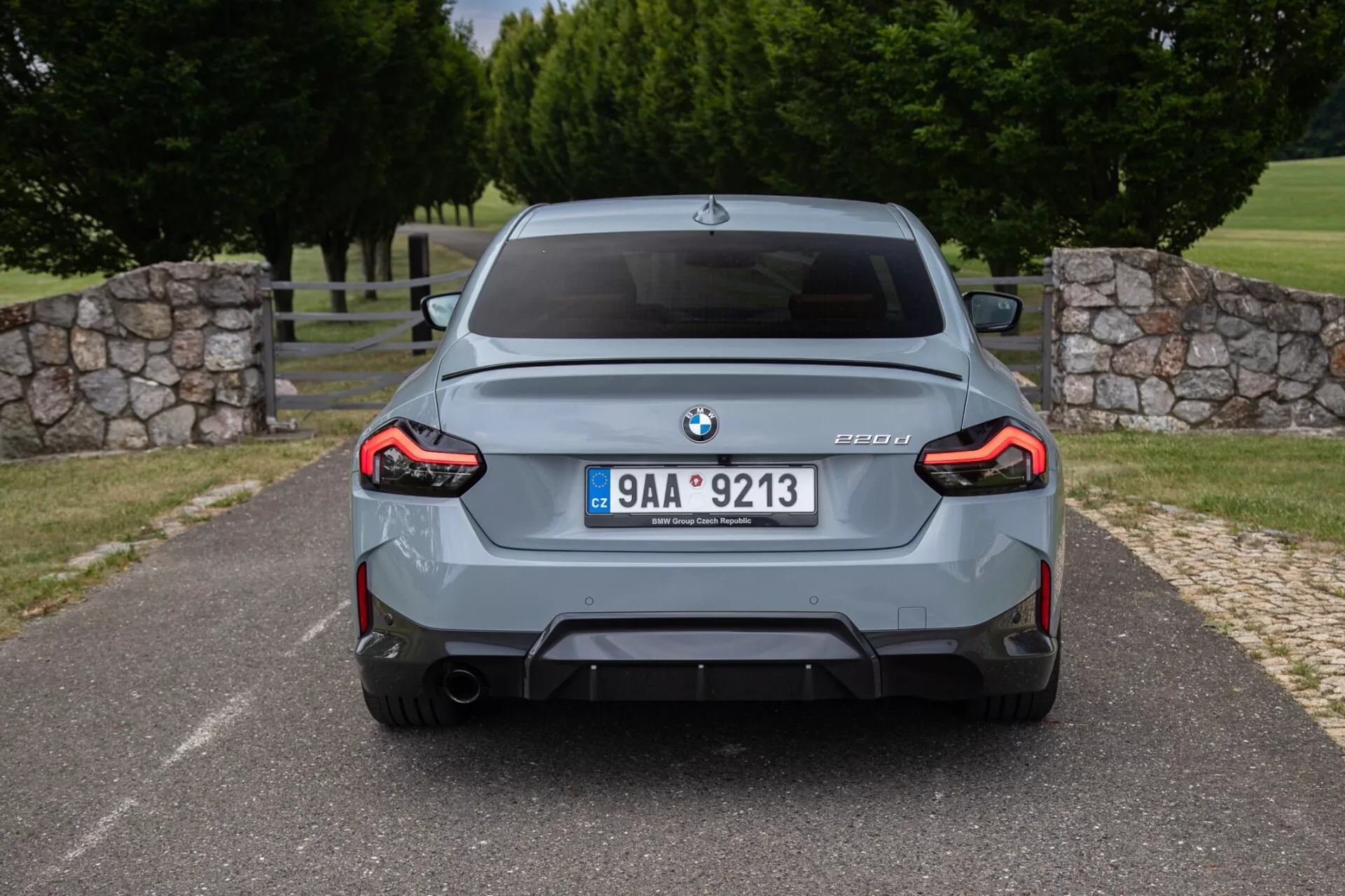 Zadný pohľad na šedý sedan BMW M3 (model G80) so značkou BMW, označením modelu M3 s informáciami o modeli G80, stredovou poznávacou značkou, športovým zadným nárazníkom s integrovaným difúzorom, dvojitými výfukmi, celoplošnými L-tvarovanými LED zadnými svetlami a rozšírenými blatníkmi.