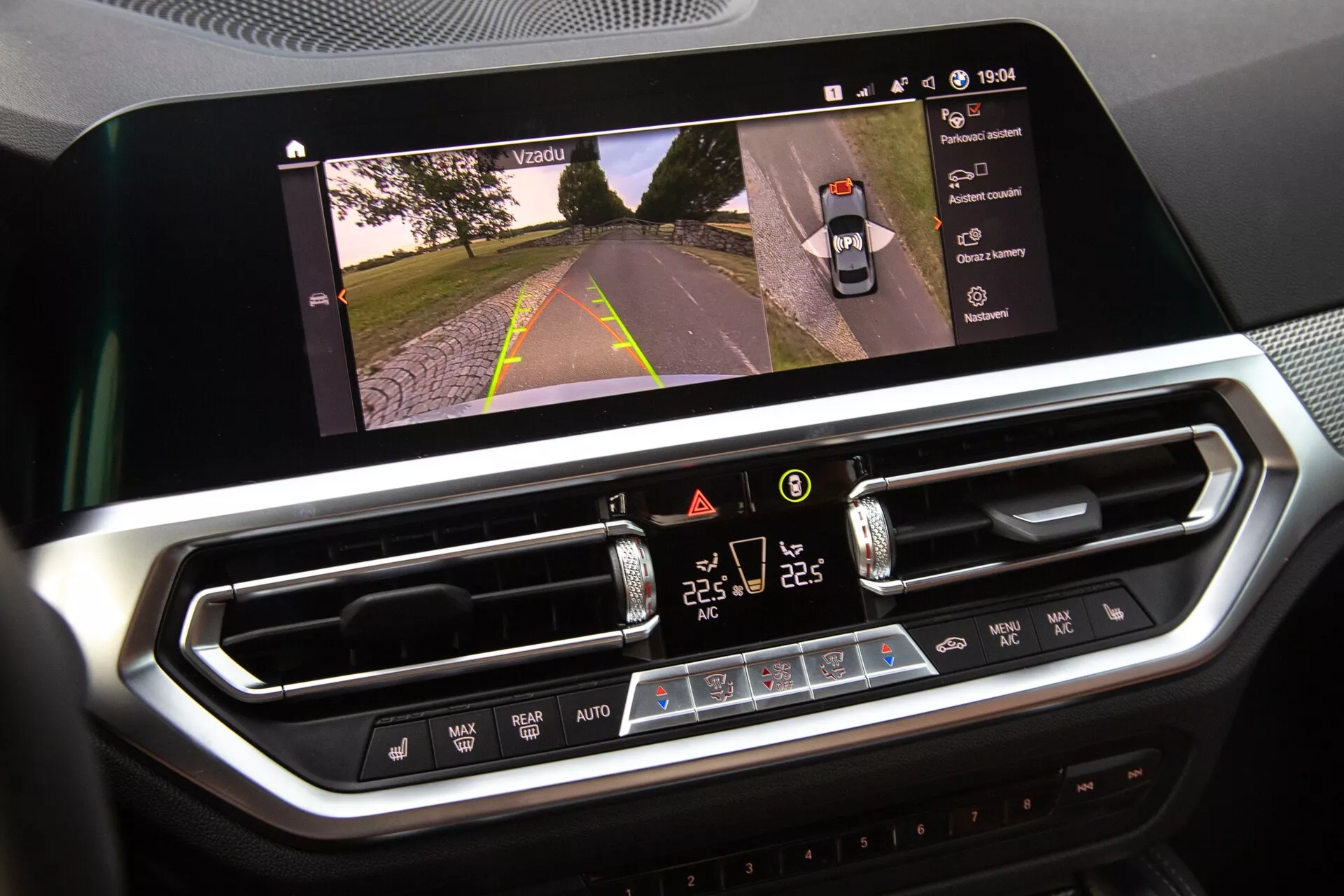 Detailný záber interiéru BMW 2er-Reihe 2022, ktorý zobrazuje infotainment displej a systém klimatizácie. Veľký, horizontálne orientovaný dotykový displej zobrazuje výhľad na cestu, čo naznačuje aktiváciu kamery, zatiaľ čo digitálne ovládanie klimatizácie nižšie obsahuje nastavenia teploty pre dve zóny, rýchlosť ventilátora a fyzické tlačidlá. Moderný dizajn s chrómovými prvkami naznačuje prémiové materiály.
