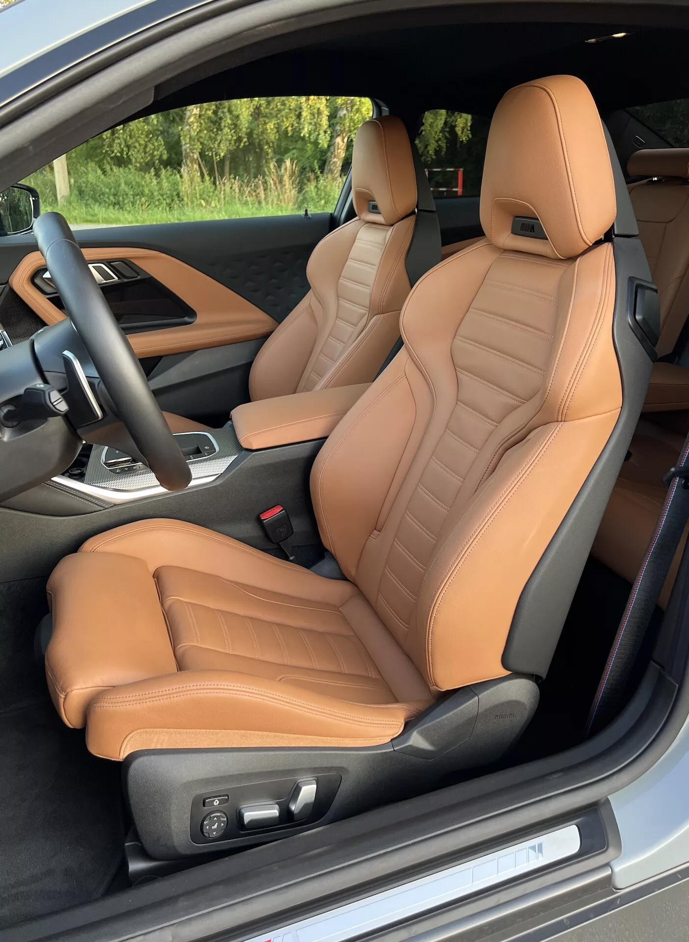 Pohľad na interiér kupé BMW série 2 z roku 2022 z pohľadu šoférových dverí, zvýrazňujúci predné sedadlá s tan koženým poťahom so štepovanými detailmi a ergonomickou podporou. Elektrické nastavenie sedadiel, čierny volant s ovládacími prvkami a čiastočný pohľad na stredovú konzolu s radiacou pákou naznačujú luxusné a výkonné vlastnosti.