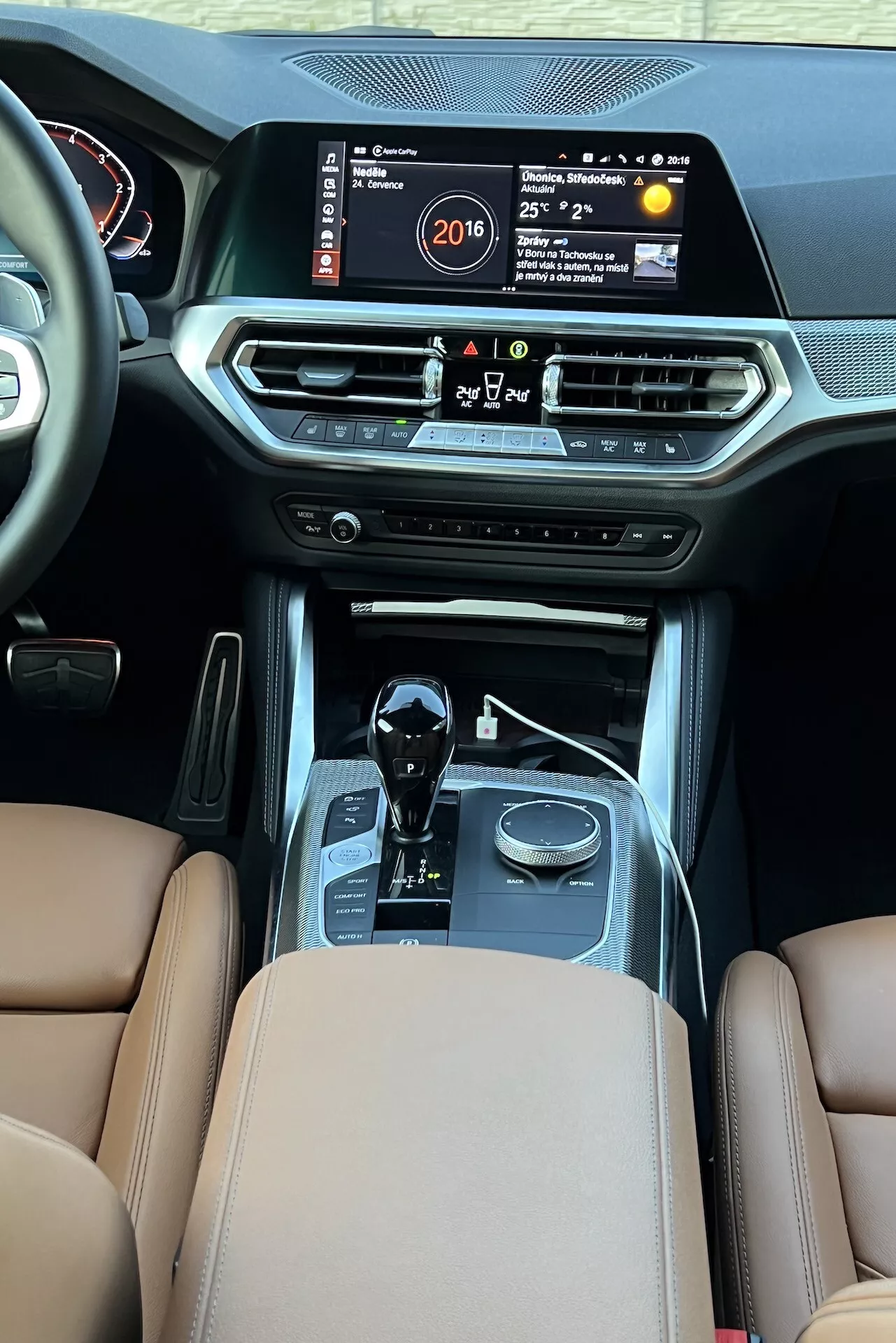 Pohľad na interiér BMW 2er-Reihe 2022 z zadnej oblasti pre pasažierov smerom k prednej palubnej doske, zvýrazňujúci stredovú konzolu s veľkou infotainment obrazovkou zobrazujúcou čas, teplotu a multimediálny systém. Funkcie zahŕňajú vetracie otvory klimatizácie, tlačidlá ovládania klímy, tlačidlo štart/stop, radiacu páku automatickej prevodovky s chrómovanými akcentmi, voliče jazdných režimov, hnedé kožené sedadlá s prémiovým čalúnením a švami, čo naznačuje luxus a pokročilú automobilovú technológiu.