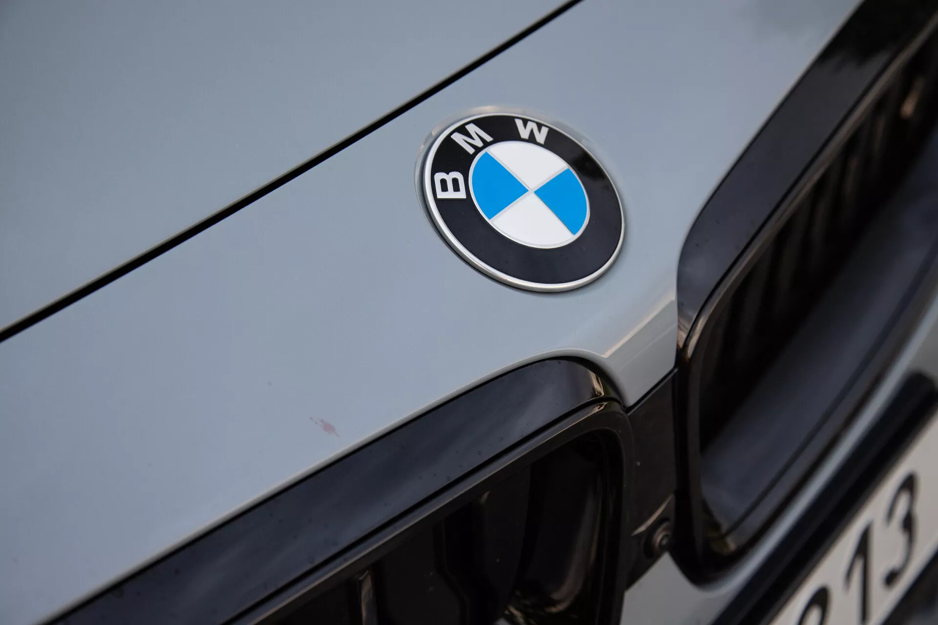 Detailný záber prednej časti BMW 2-Series Coupe z roku 2022, s výrazne umiestneným čierno-modro-bielym znakom BMW na kapote. Obrázok zachytáva horný okraj ikonickej obličkovej mriežky v pozadí, s kovovým strieborným alebo svetlo šedým exteriérom odrážajúcim okolité svetlo. Ostrá zameranosť na znak zdôrazňuje luxusnú značku a dobre udržiavaný povrch vozidla.
