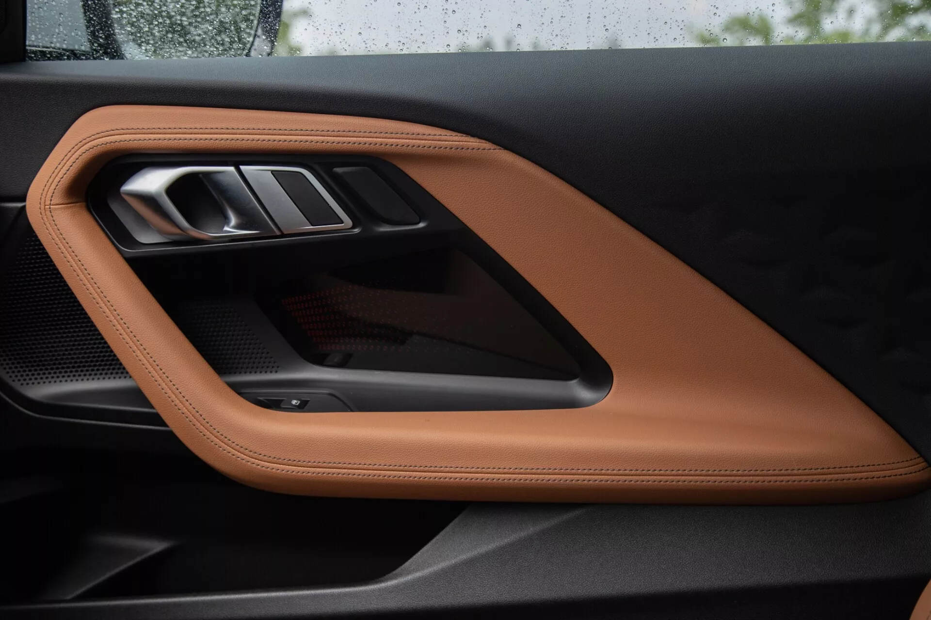 Detailný pohľad na vnútorný dverový panel BMW 2 Series Coupe 2022, zobrazujúci luxusne prešívanú kožu farby tan alebo koňakovú, elegantnú kovovú kľučku dverí a športový karbónový akcent, zdôrazňujúci vysoko kvalitné materiály a sofistikované dizajnérske prvky.