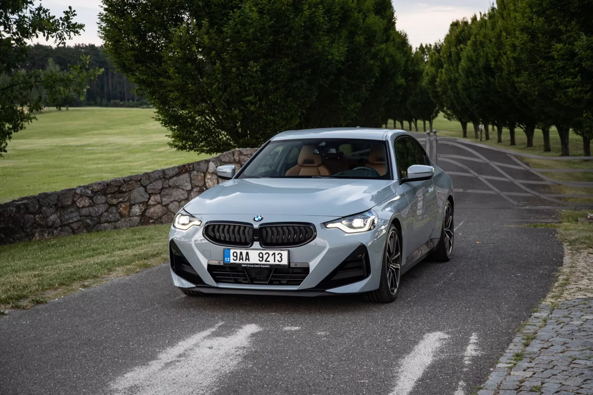 Podrobný obrázok kupé BMW radu 2 z roku 2022 zachytený z trojštvrťového predného uhla, ktorý ukazuje jeho predné a bočné profily na pokojnej asfaltovej ceste obklopenej upravenými stromami a kamenným múrom. Výrazná mriežka BMW a elegantné LED svetlomety zdobia prednú časť, zatiaľ čo prívody vzduchu na nárazníku zvyšujú jeho športový vzhľad. Kontúrované línie kapoty naznačujú výkon, dobre definované blatníky naznačujú silu a viacramenné zliatinové kolesá signalizujú športovú úpravu. Bočný profil má charakterovú líniu pre pocit pohybu, bočné zrkadlá s integrovanými smerovkami a náznak strešného okna. Uhol čelného skla a čisté bočné okná pridávajú vyvážený vzhľad. Mäkké osvetlenie, pravdepodobne skoro ráno alebo neskoro popoludní, vytvára jemné tiene a čistú prezentáciu dizajnu a farby vozidla.