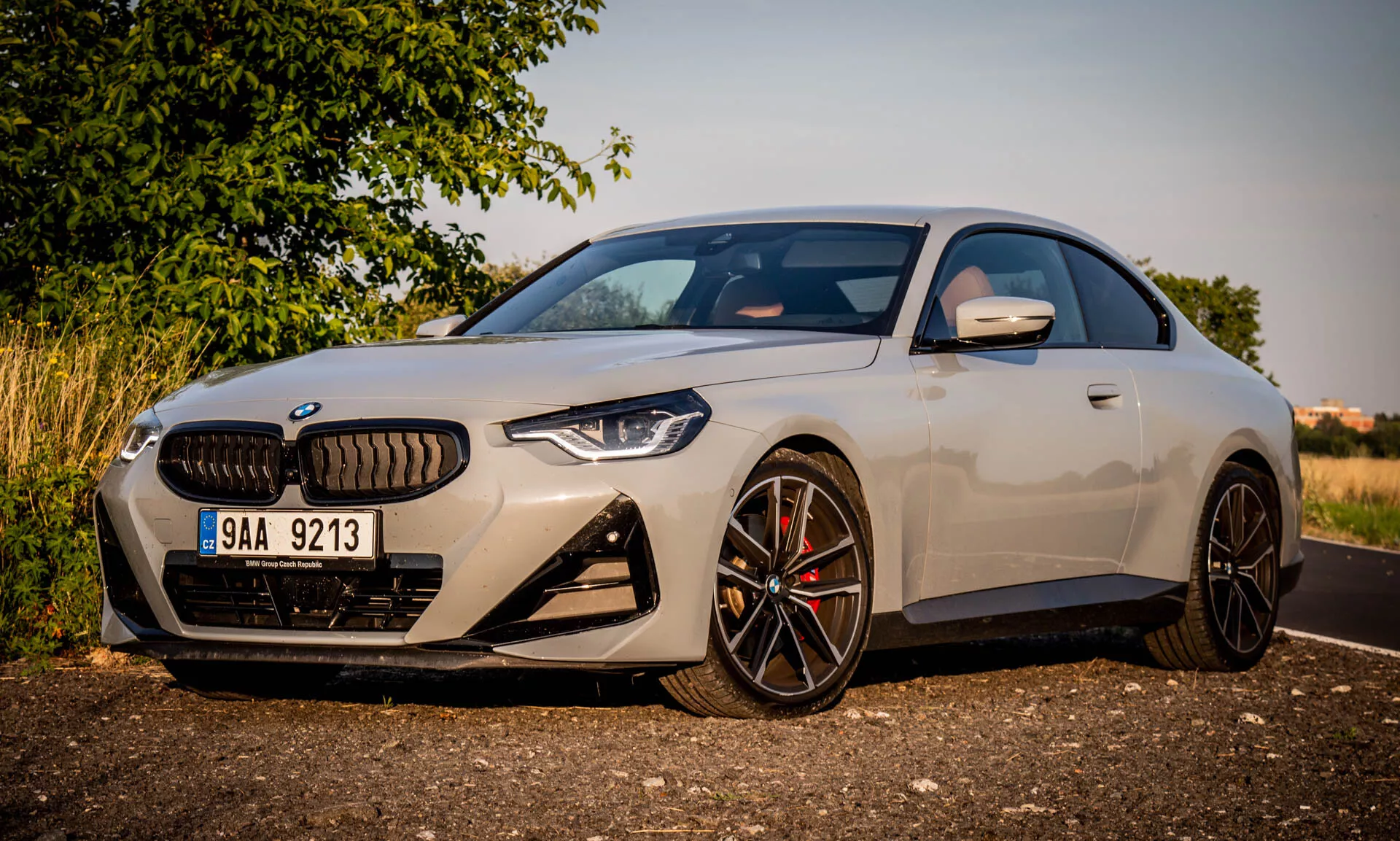Obrázok BMW radu 2 kupé z roku 2022. Moderné luxusné vozidlo je zaparkované pod uhlom, zvýrazňujúce prednú ľavú stranu počas zlatej hodiny. Záber sa sústreďuje na výrazné dizajnové prvky ako veľká mriežka v tvare obličky, hranaté LED svetlomety, športový predný nárazník s veľkými prieduchmi a viaclúčové čierne zliatinové kolesá s vysoko výkonnými brzdovými strmeňmi. Aerodynamické bočné zrkadlo má integrované smerovky. Elegantná karoséria a prvotriedna farba zdôrazňujú kompaktný, športový profil vozidla a nízku líniu strechy.