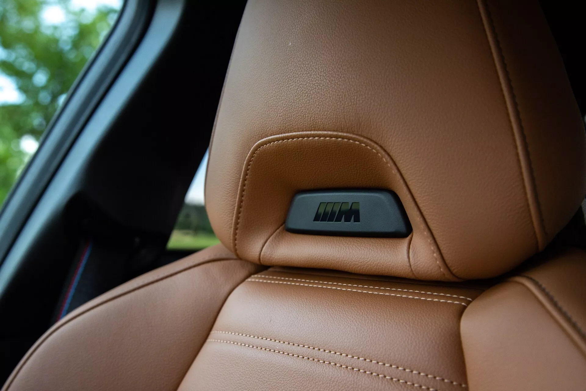 Detailný pohľad na hornú časť hnedého koženého sedadla v BMW série 2 Coupé 2022, zdôrazňujúci presné viditeľné švy a ergonomicky navrhnutý obrys pre pohodlie a podporu. Na hornej časti sedadla je vpredu zabudovaný odznak, ktorý označuje technologické funkcie, ako sú elektronické nastavenia alebo funkcie vyhrievania. Záber je urobený z mierne nižšieho uhla, zvýrazňuje luxusnú textúru kože a rozmazáva pozadie, ktoré zahŕňa časť okna auta.