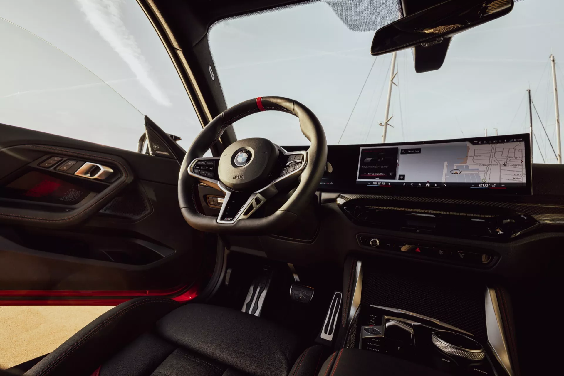 Pohľad do interiéru BMW 2er-reihe 2024 (G42-G87 Facelift) z miesta spolujazdca. Na obrázku je trojramenný volant s integrovanými ovládacími prvkami, digitálny prístrojový panel, veľký infotainment displej a minimalistické ovládanie klimatizácie. Viditeľné červené ambientné osvetlenie a automatická prevodovka v stredovej konzole. Panel dverí obsahuje ovládanie okien a zrkadiel, rovnako ako mriežku reproduktora. Kožené sedadlá s kontrastným prešívaním pridávajú na luxusnom dojme.