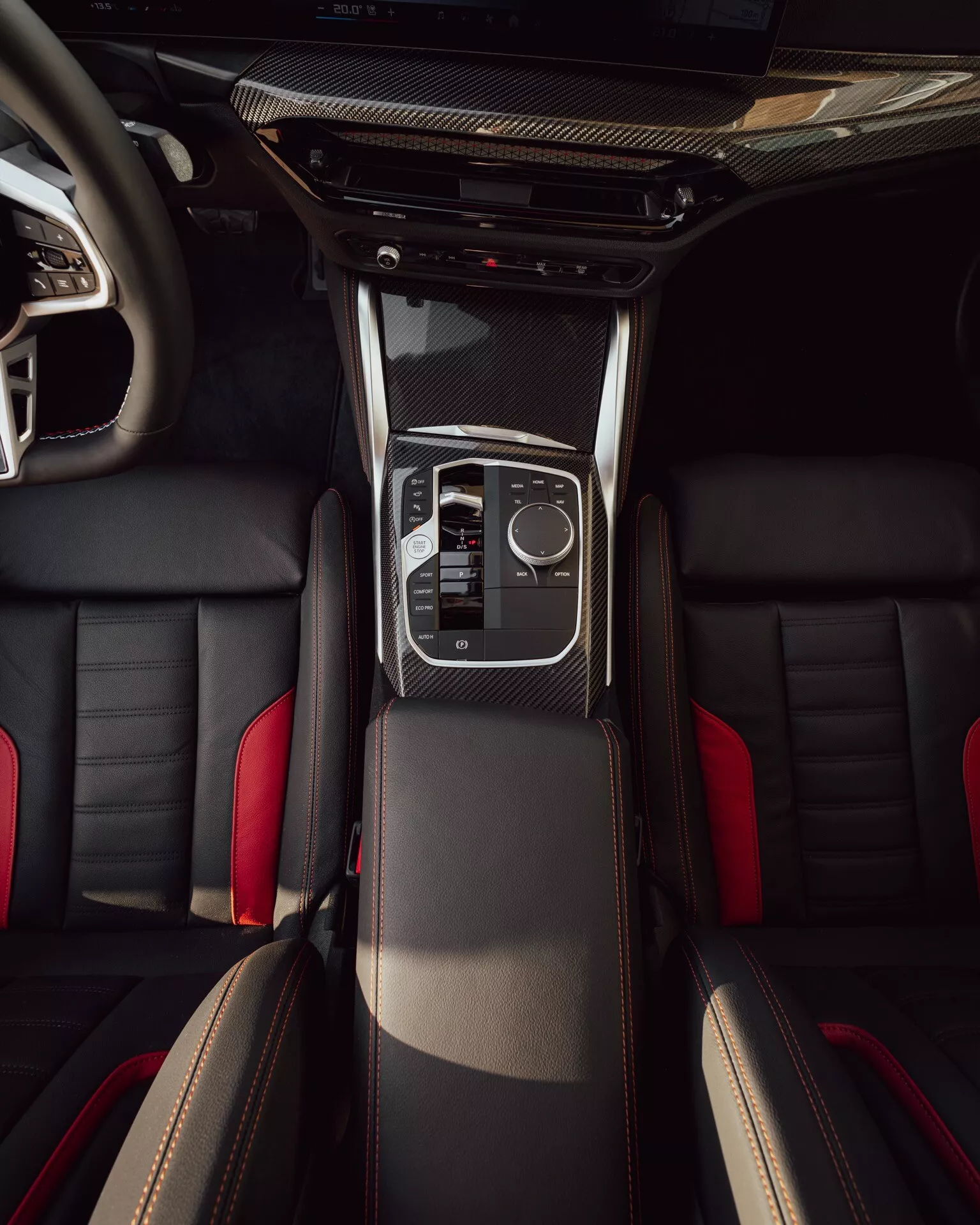 Pohľad na interiér BMW radu 2 2024 (facelift G42-G87) z vtáčej perspektívy, ukazujúci stredovú konzolu s karbonovým výbavou, elegantnú radiacu páku, elektronický touchpad, klimatické ovládače, športové kožené predné sedadlá s kontrastným šitím a čiastočný pohľad na zadné sedadlá.