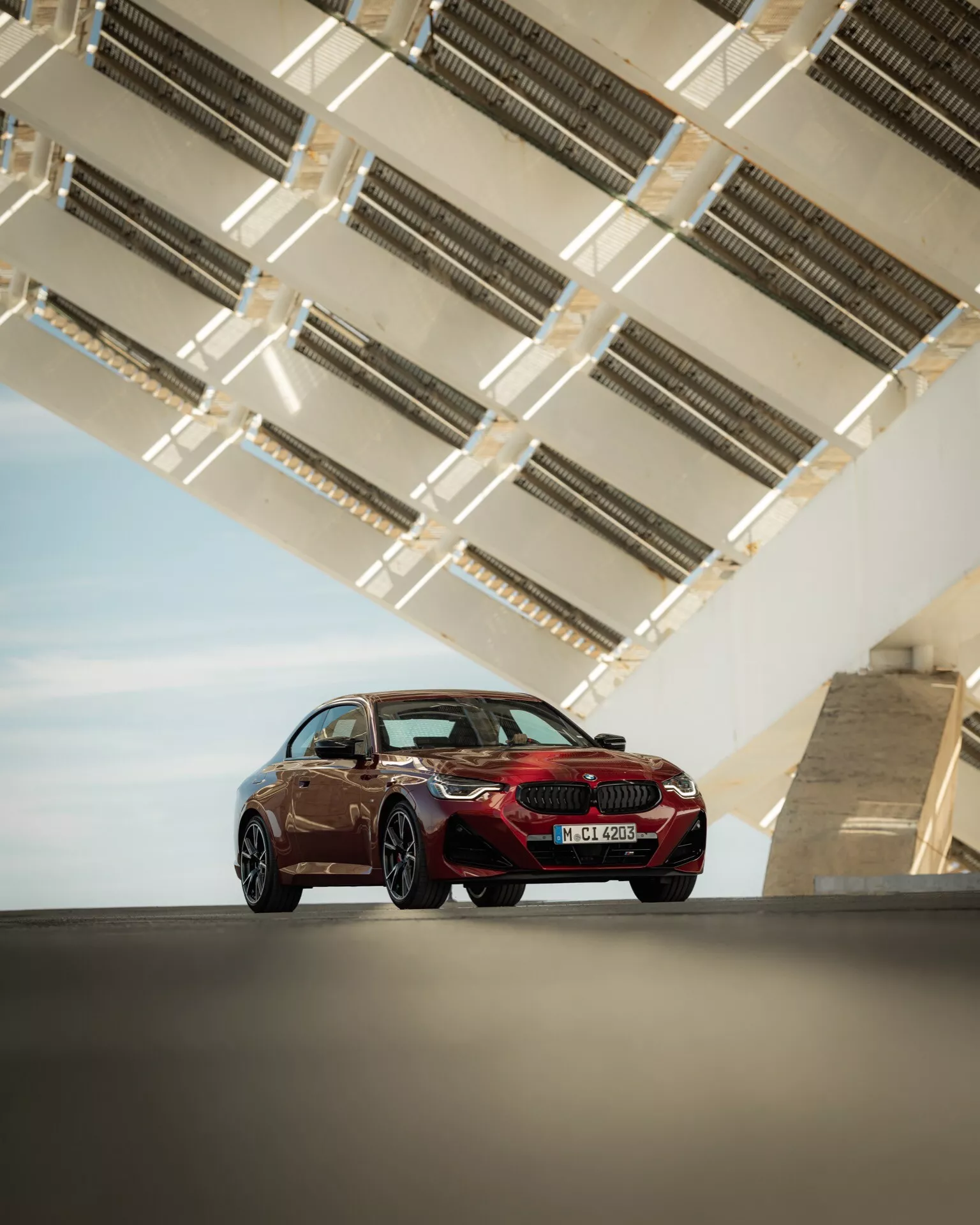 Obrázok zobrazuje luxusný športový sedan BMW radu 2 z roku 2024 (facelift G42-G87) z nízkeho uhla, zvýrazňujúc jeho postavu a dizajnové línie. Záber urobený z predného trojštvrťového pohľadu zobrazuje stranu vodiča auta, ukazuje prednú mriežku, LED svetlá, obrys kapoty, bočný profil, kolesá a dvere. Sedan je umiestnený na elegantnej vydláždenej ploche s modernou geometrickou architektonickou konštrukciou nad ním, pravdepodobne časť mostu alebo zvýšenej diaľnice, s kovovými prvkami zdôrazňujúcimi dizajn a farbu vozidla. Aerodynamická silueta, športové ráfiky s veľkým priemerom a aerodynamicky tvarované vonkajšie zrkadlá naznačujú pokročilú aerodynamiku a modernú technológiu vozidla.