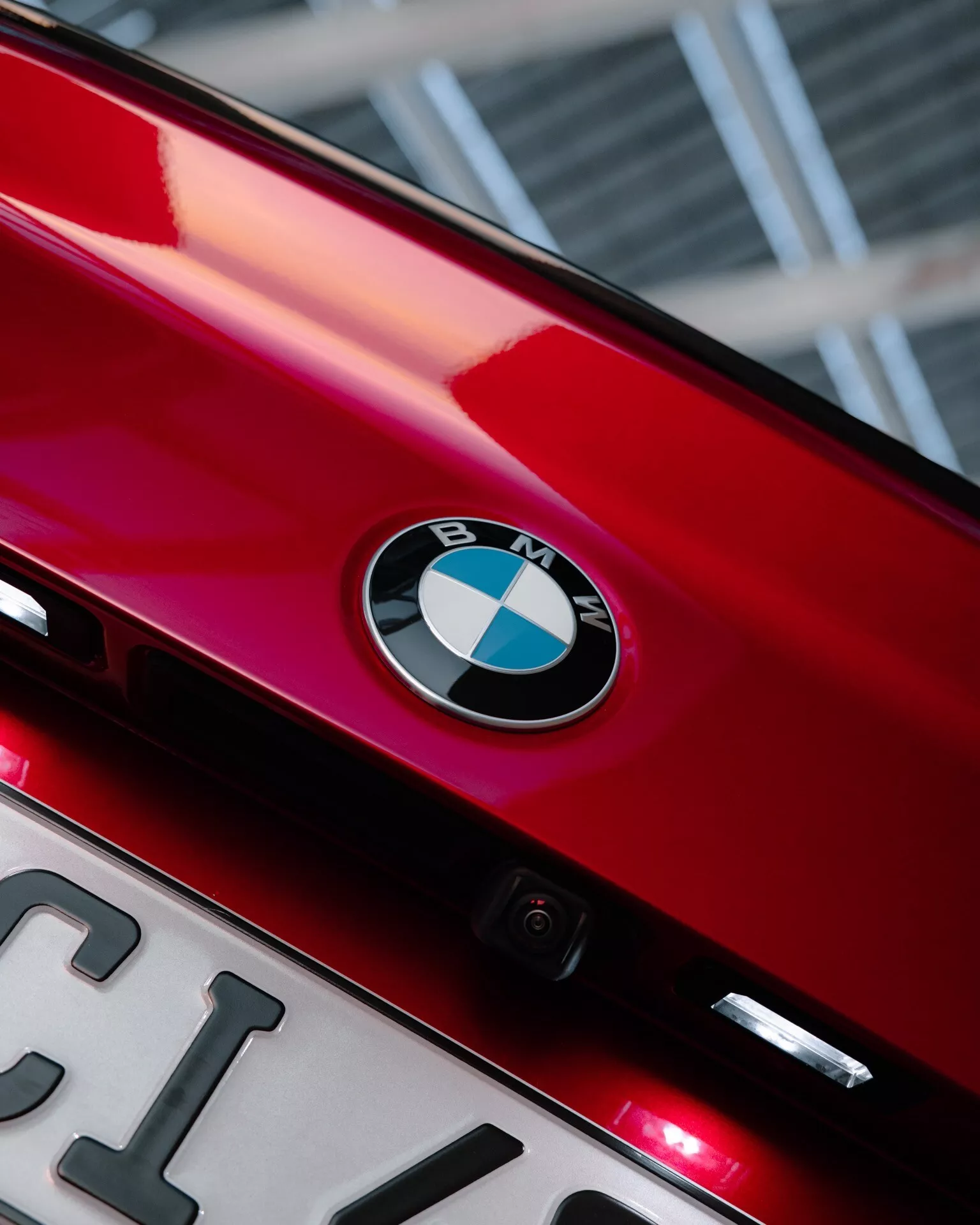 Detailný pohľad na zadnú časť lesklého červeného BMW radu 2 z roku 2024 (G42-G87 facelift), ukazujúci centrálny znak BMW na veku kufra. Pod znakom je čiastočne viditeľná evidenčná tabuľka a malá zadná kamera zdôrazňujúca technologické prvky. Dve malé červené svetlá pod znakom môžu byť doplnkové brzdové svetlá alebo zadné hmlové svetlá. Uhol fotografie smerom nahor a tesný rám zvýrazňujú identitu značky a elegantné dizajnové prvky s bokeh efektom na pozadí.