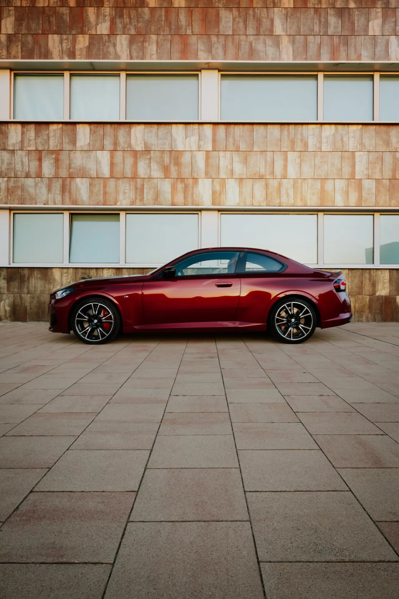 Moderný luxusný sedan hlbokej červenej farby BMW 2er-Reihe 2024 (G42-G87 Facelift) zaparkovaný na veľkej dlažbe s pozadím architektonickej fasády farby hrdze, bočný profil, elegantné línie, chrómové okenné lišty, športové hliníkové kolesá, prirodzené osvetlenie.