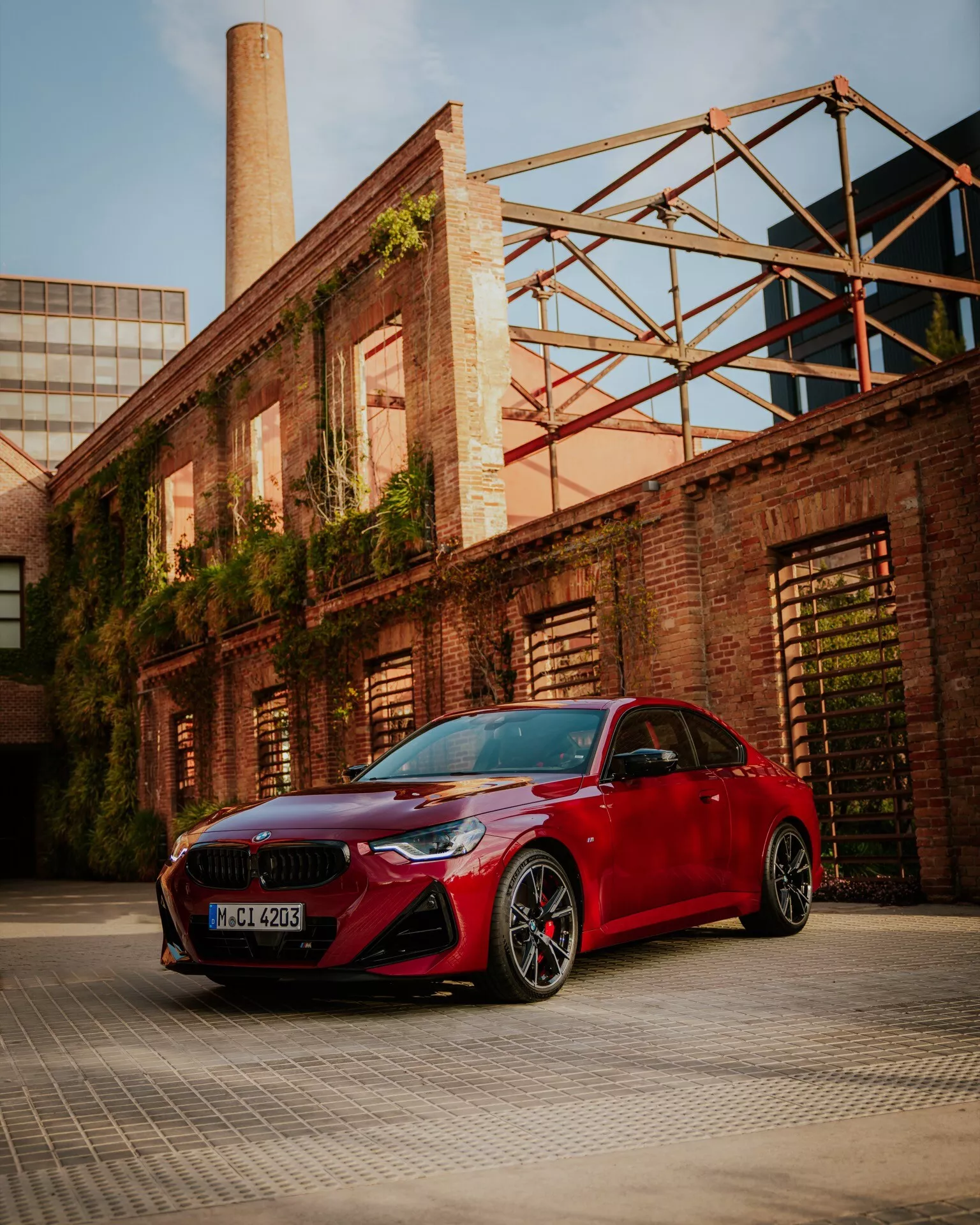 Nízky uhol záberu na červené BMW radu 2 2024 (G42-G87-facelift) zaparkované v priemyselnej alebo skladovej časti, ktorý ukazuje jeho športový predný nárazník s veľkými vstupmi vzduchu, úzkymi elegantnými svetlometmi, mriežkou v tvare obličiek a aerodynamickými bočnými zrkadlami. Obrázok tiež obsahuje viaclúčové čierne zliatinové kolesá s nízkoprofilovými pneumatikami a viditeľné brzdové strmene. Sochárske línie na kapote a lesklý lak sú zvýraznené teplým okolným svetlom, zdôrazňujúc luxusné a výkonné dizajnové prvky auta.