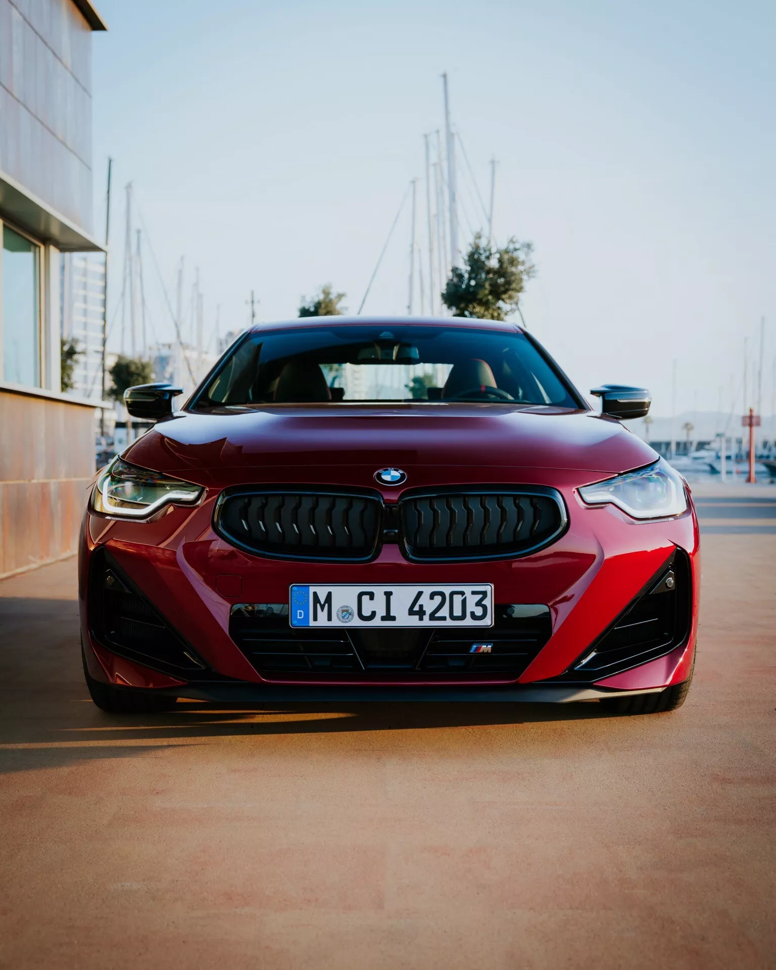 Predný pohľad na červené auto BMW 2er-Reihe 2024 (G42-G87 Facelift) zachytený z nízkeho uhla, kde vyniká symetrická predná časť. Výrazná mriežka BMW s jemným čiernym sieťovým dizajnom a modro-bielym emblémom je lemovaná ostrými, uhlovými LED svetlometmi. Aerodynamický predný nárazník s veľkými prívodmi vzduchu a čiernymi vložkami zvyšujú športový vzhľad. Rozmazané pozadie s rahnami naznačuje prostredie pri maríne alebo dokoch.